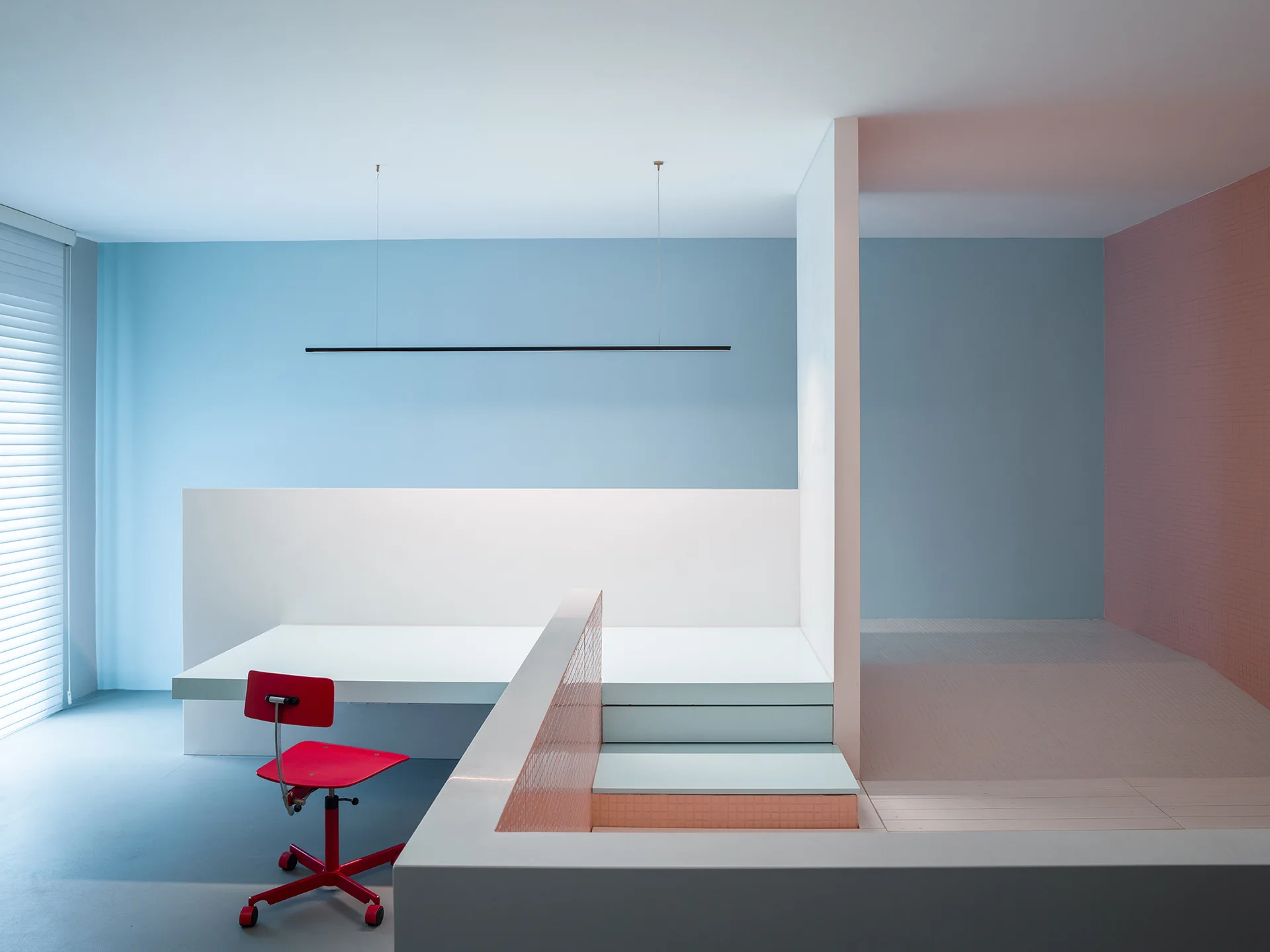 A minimalist home office features a white built-in desk, a red chair, a blue painted wall, and steps leading to a raised platform.