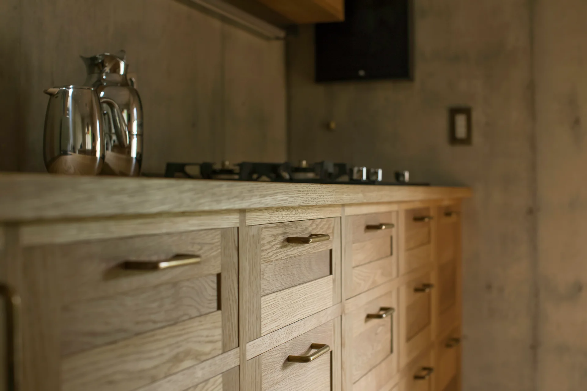 Close-up of light wood kitchen drawers with brass handles beneath a wooden countertop, set against a textured concrete wall.