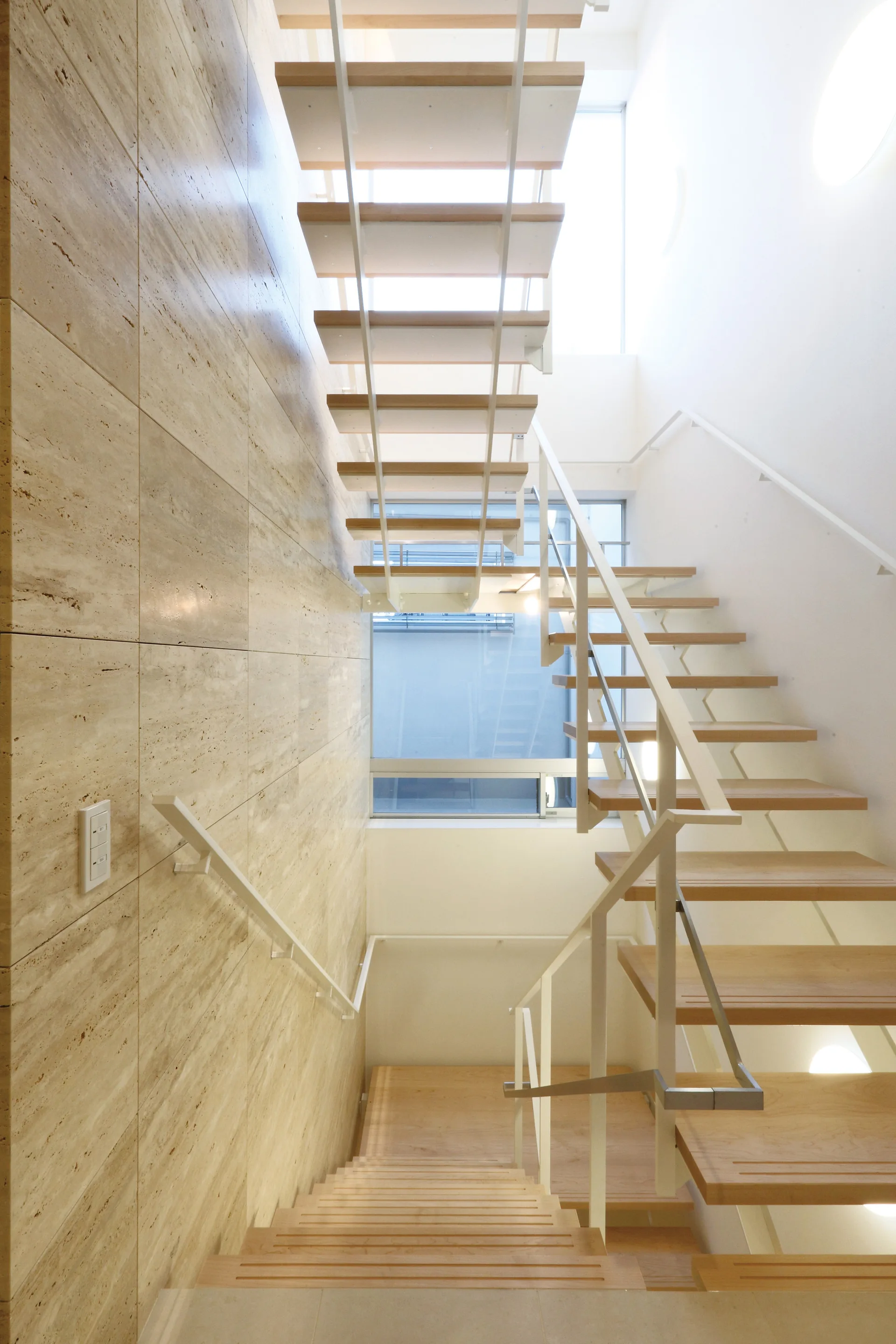 A minimalist open-riser wooden staircase with a white metal railing is positioned next to a large-format beige travertine stone wall.