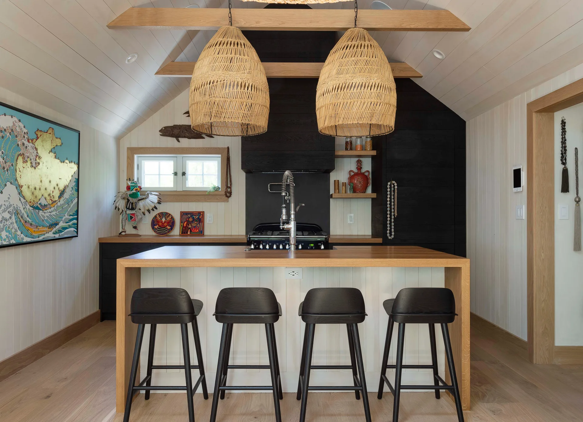 A wooden kitchen island with black bar stools is illuminated by two large woven wicker pendant lights in a room with white vertical plank walls.