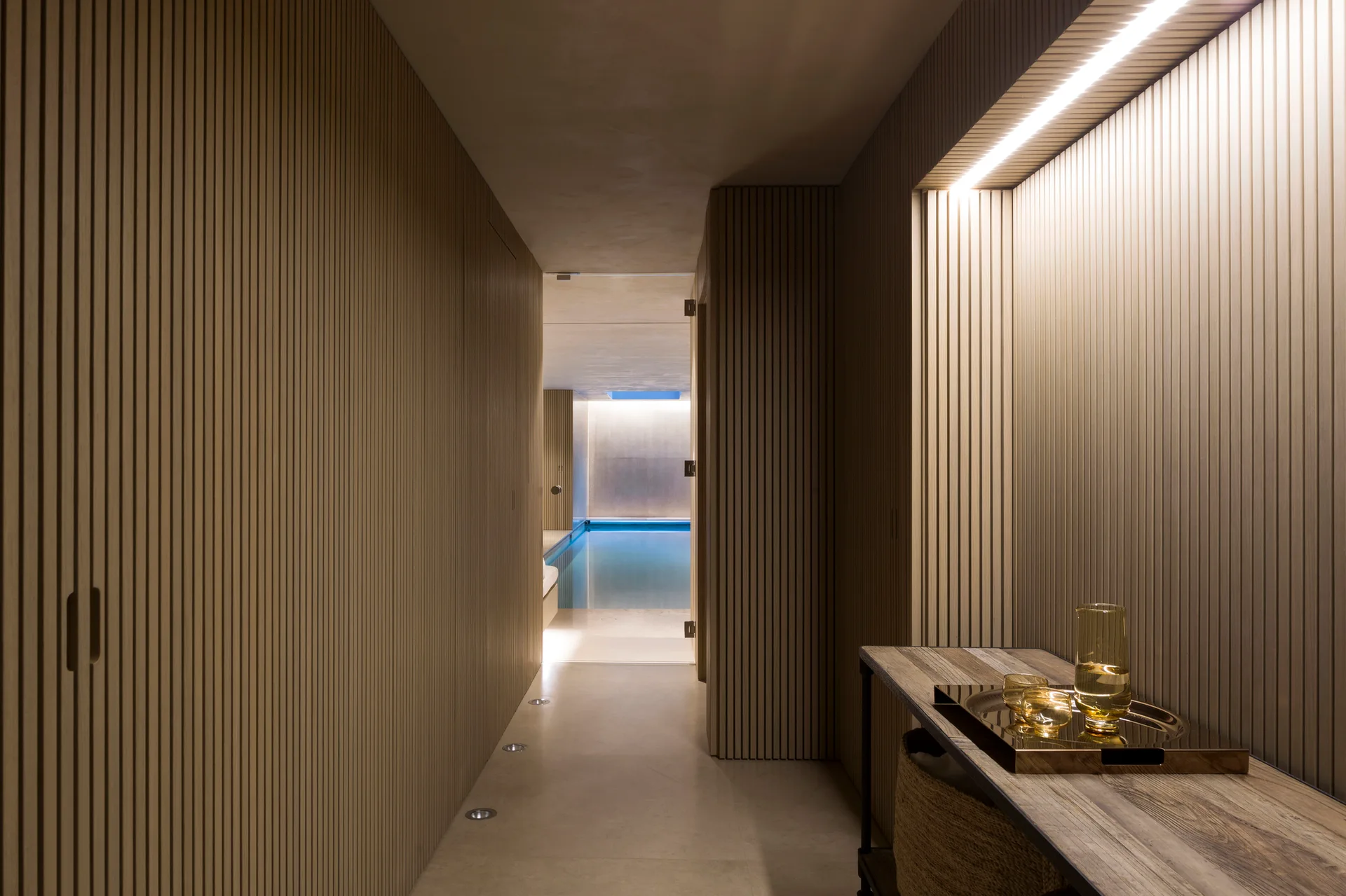 A long hallway with vertical slatted wood wall panels leads to a brightly lit swimming pool area at the far end.