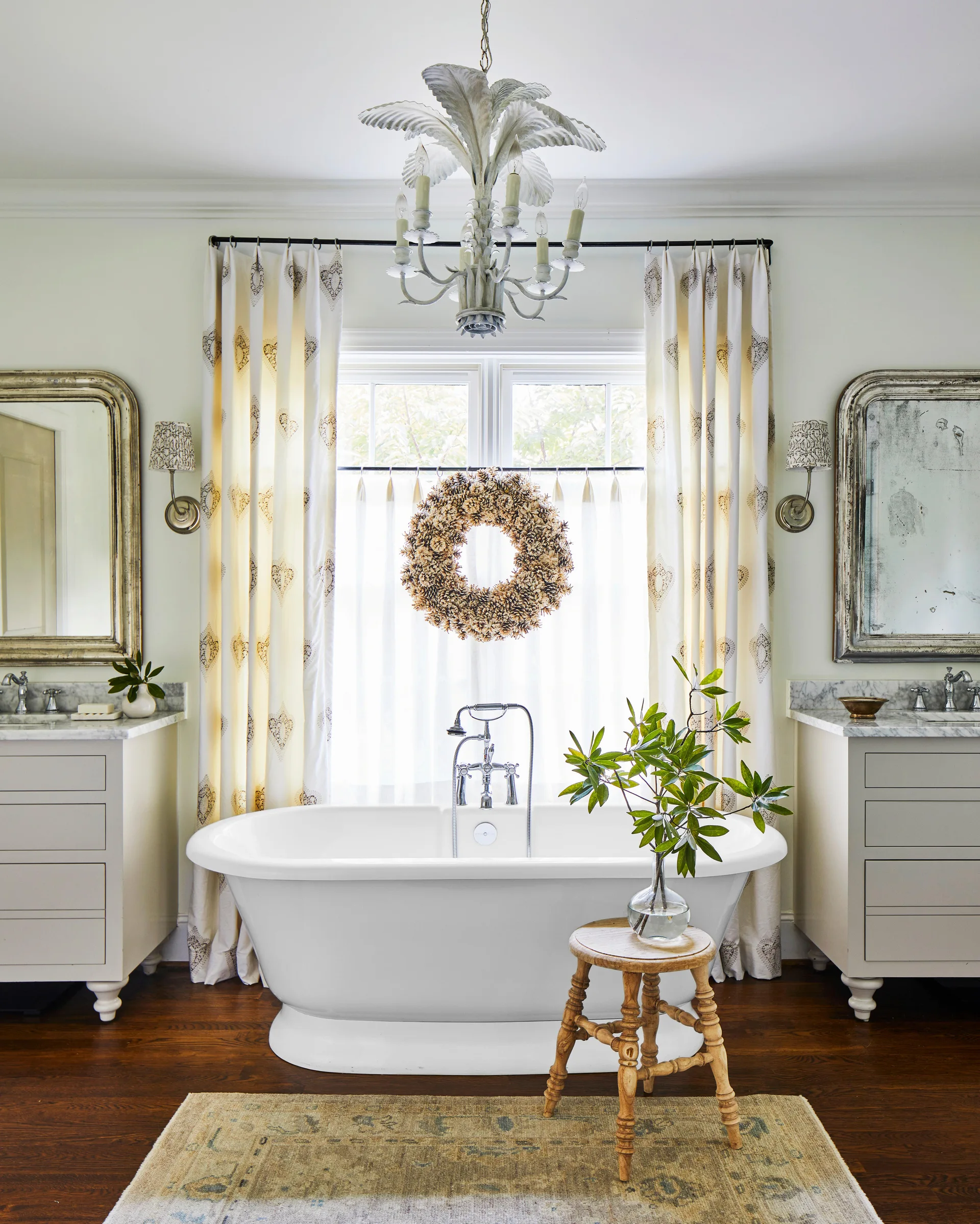 A symmetrical bathroom features a central white clawfoot tub, two matching vanities with marble tops, patterned drapes, and a palm leaf chandelier.