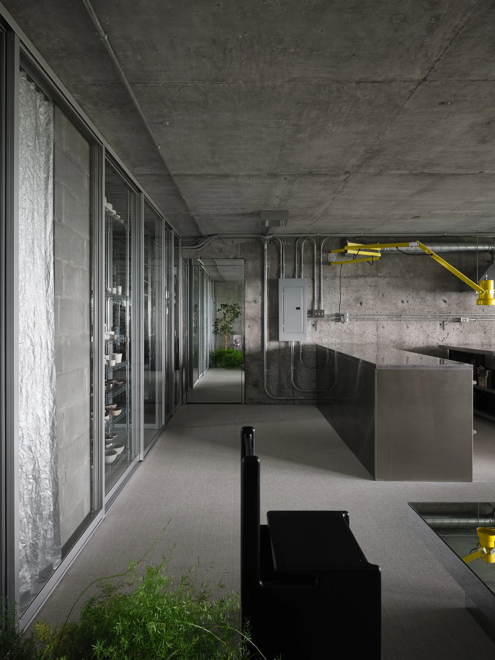 An industrial kitchen features raw concrete walls and ceiling, a large stainless steel island, glass-front cabinets, and a black minimalist chair.