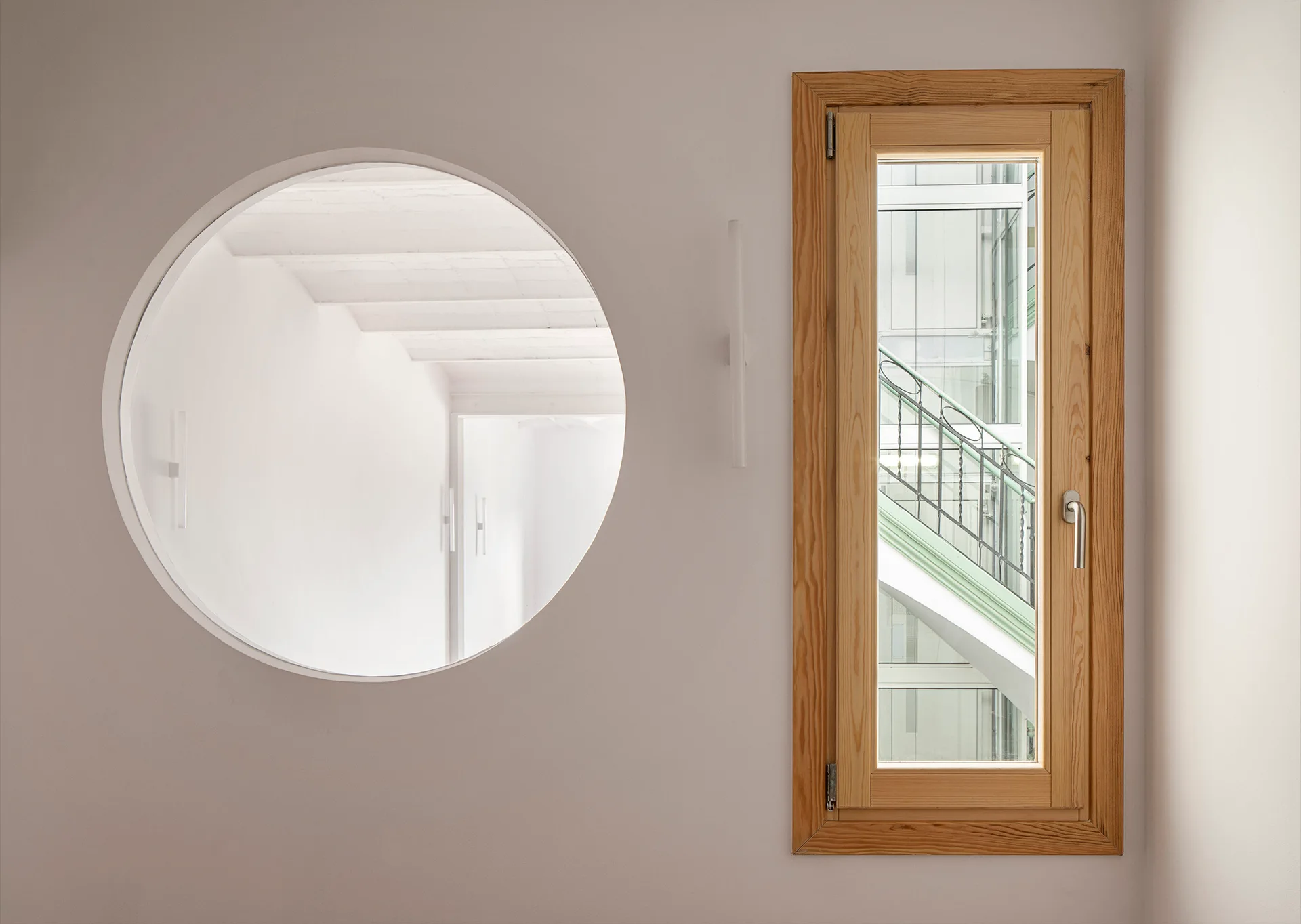 A minimalist white wall featuring a circular interior cutout looking into a beamed room alongside a narrow rectangular window framed in light wood.