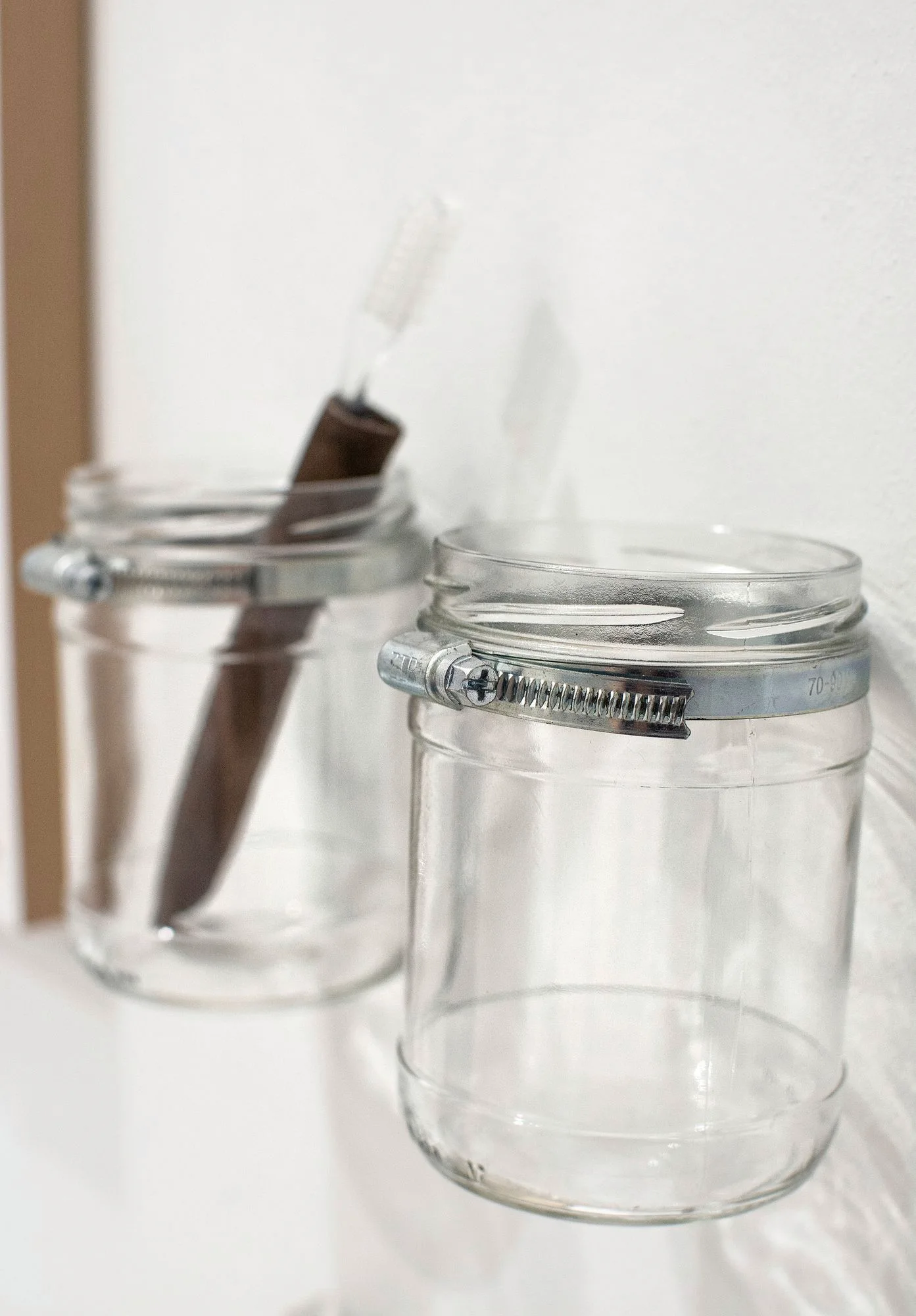 Close-up of two clear glass jars secured with silver metal hose clamps against a white wall, with one containing a brown-handled toothbrush.