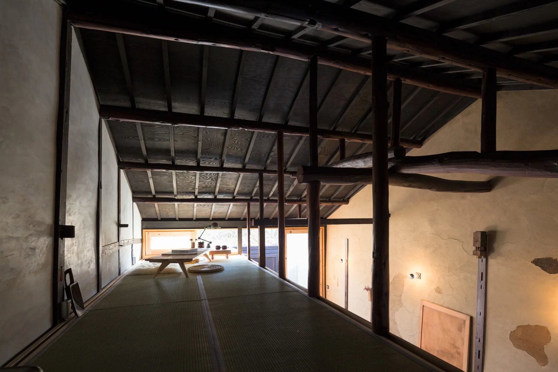 A traditional Japanese loft features tatami mat flooring, weathered plaster walls, and dark exposed timber beams under a sloped ceiling.