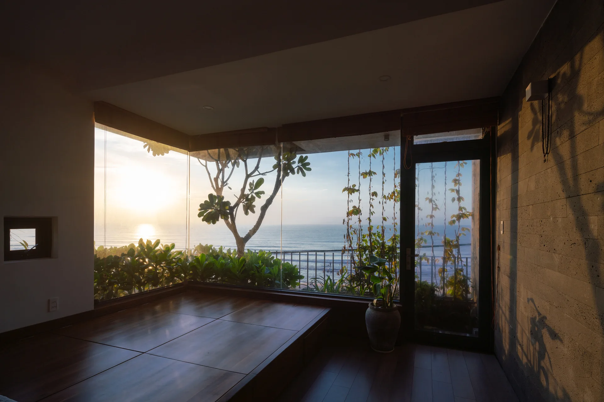 A minimalist room features a raised dark wood platform beside floor-to-ceiling corner windows looking out at a coastal ocean sunset.