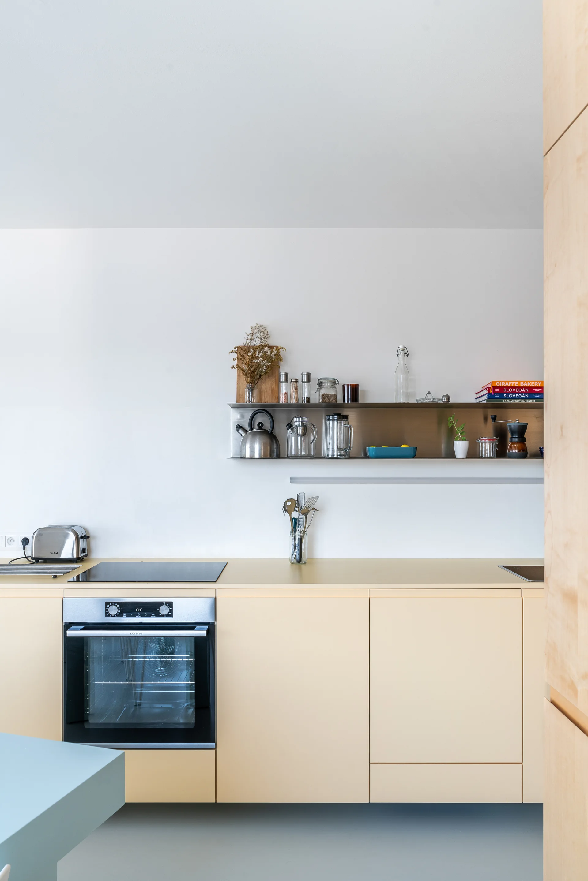 Minimalist kitchen featuring pale yellow cabinets, an integrated oven and cooktop, and a stainless steel floating shelf holding kitchenware.