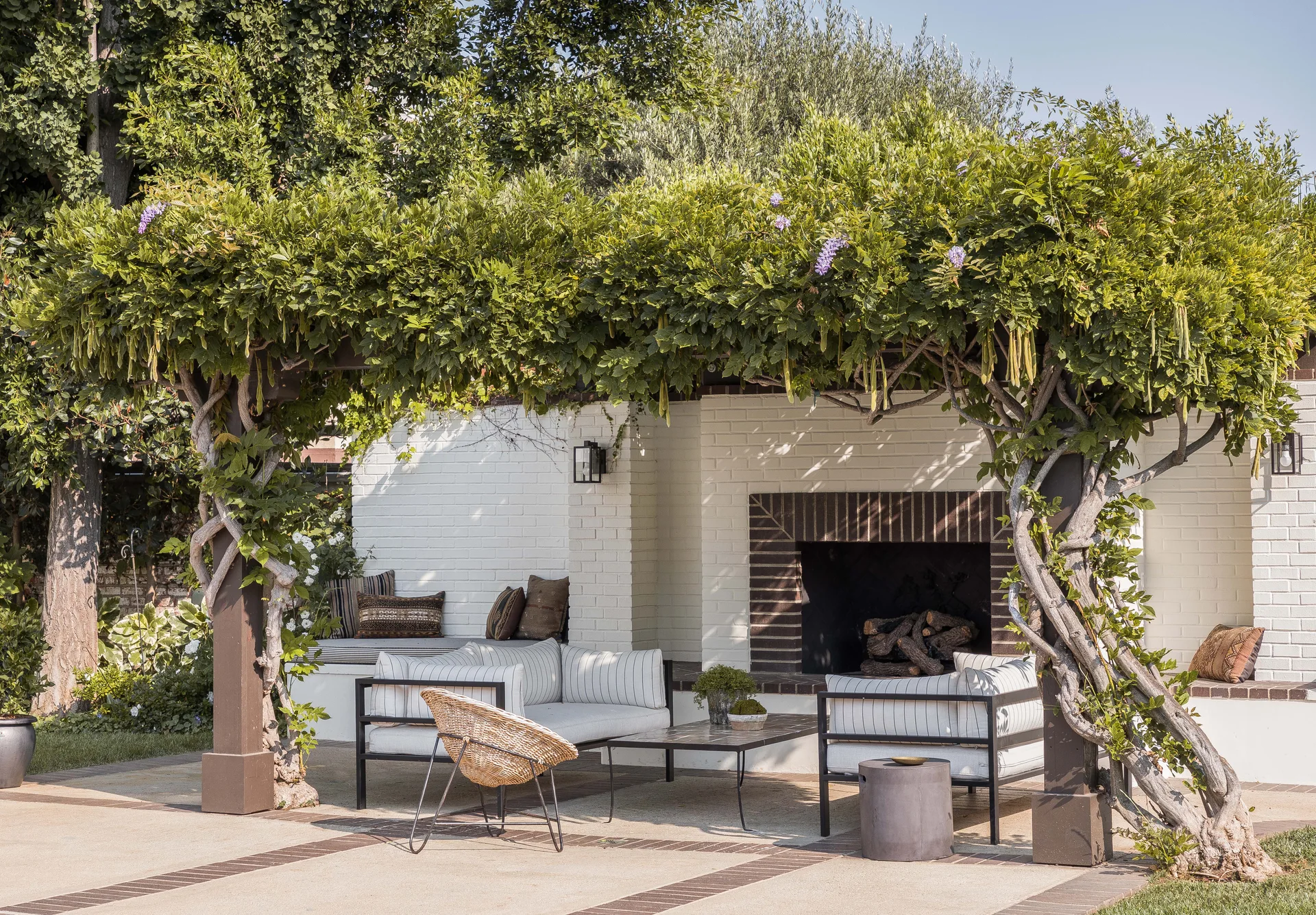 An outdoor patio features a vine-covered pergola over striped sofas, a wicker chair, and a white brick fireplace.