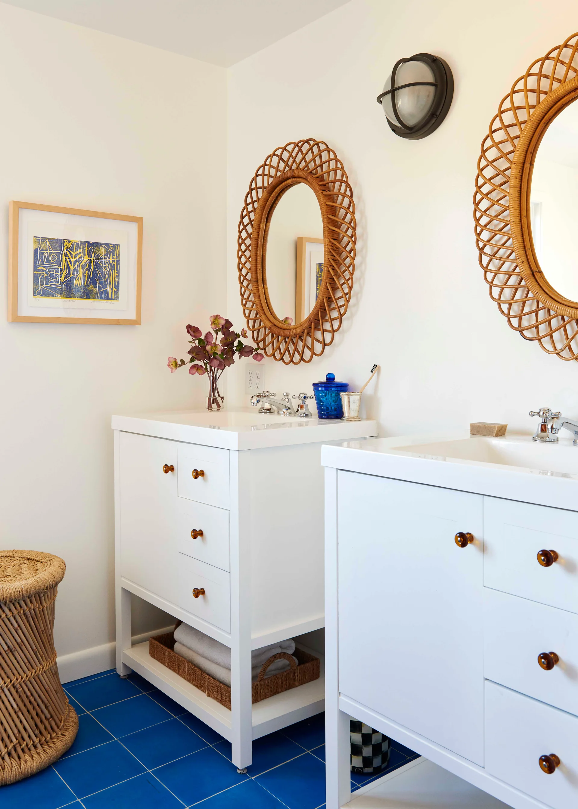 A bathroom featuring twin white vanities with chrome faucets, two round rattan mirrors, and bright blue square tiled floors.