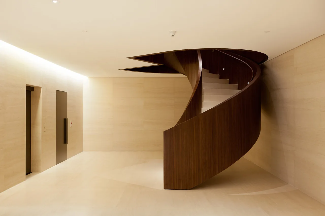 A dark wood spiral staircase curves gracefully in a minimalist foyer with beige stone walls, tiled flooring, and recessed elevator doors.