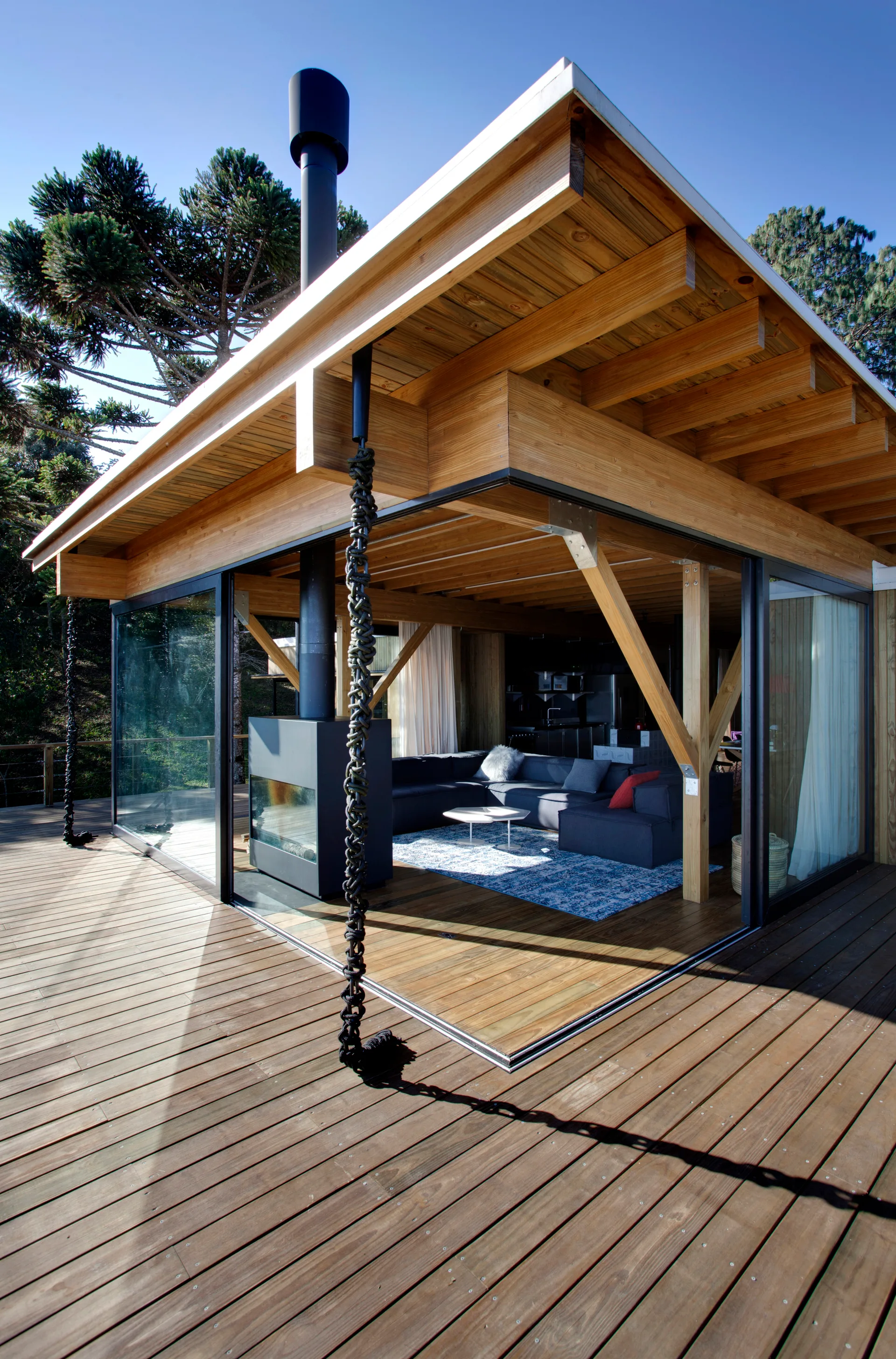 A modern timber pavilion featuring an open-corner sliding glass door system that connects an indoor living room to a wide wooden deck.