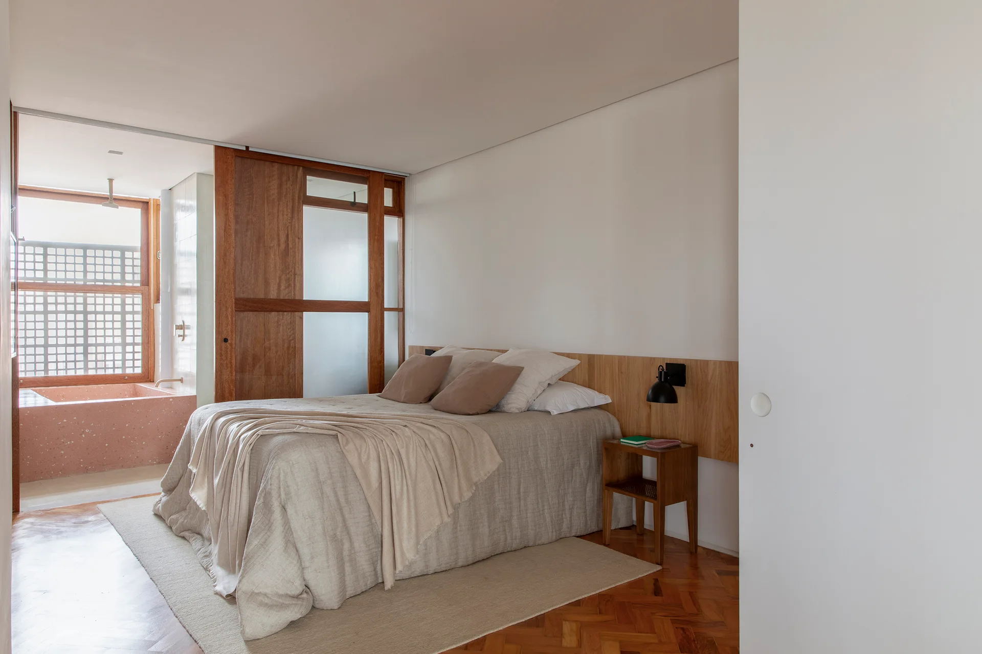 A minimalist bedroom featuring a bed with beige linens, a low wooden headboard panel, parquet flooring, and a sliding wood partition leading to a bathroom.