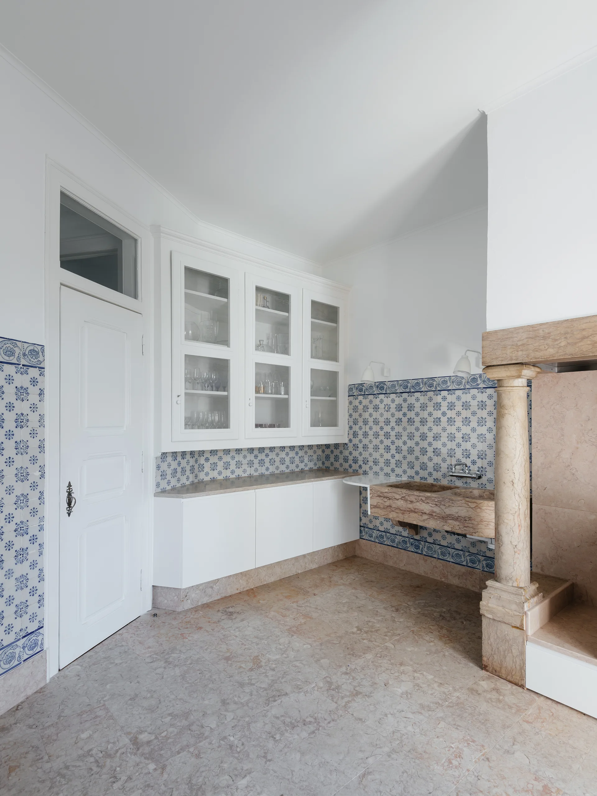 A traditional kitchen with white cabinetry, a wall-mounted carved stone sink, blue and white patterned tile walls, and a classical stone column.