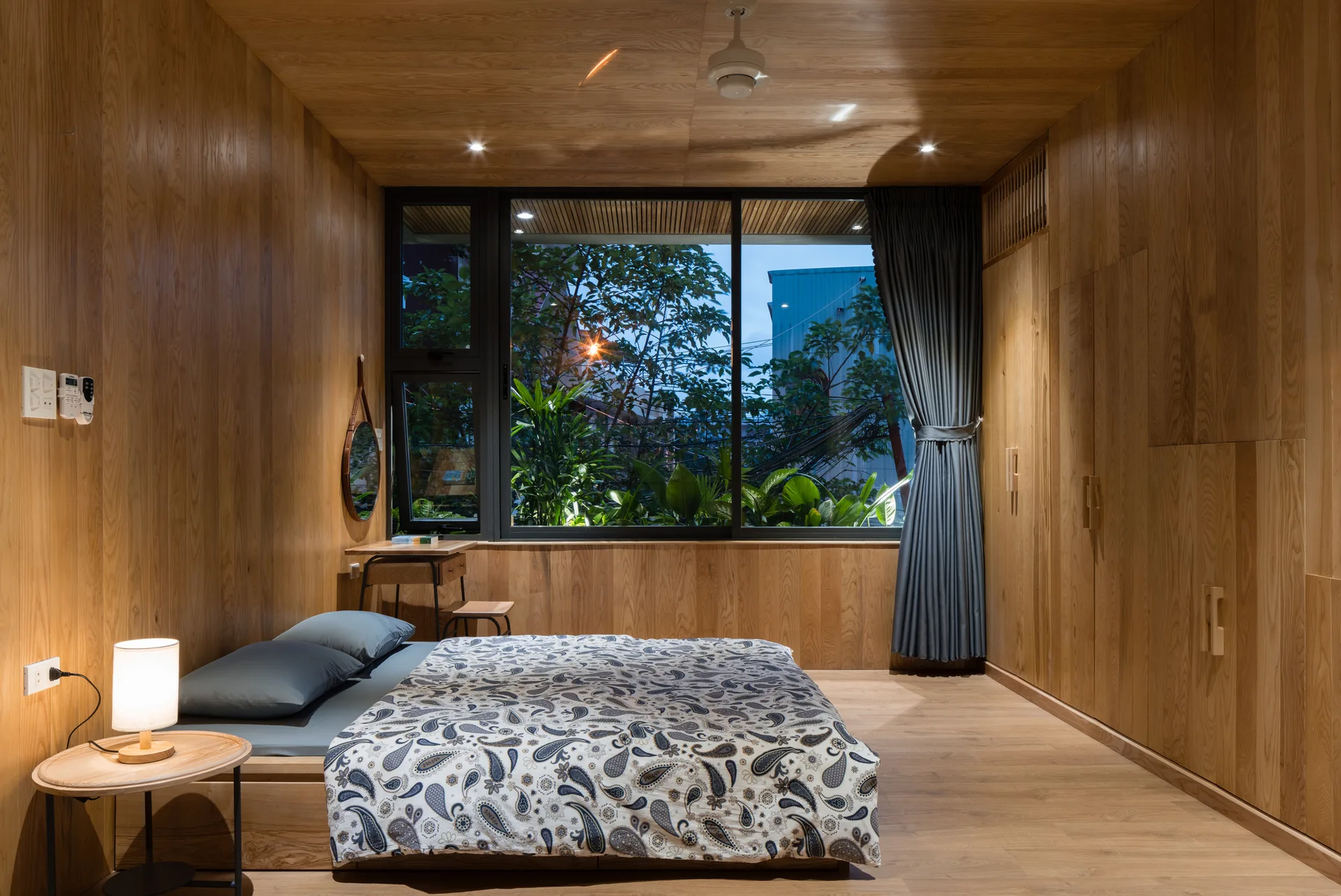A minimalist bedroom with light wood paneling on the walls and ceiling, featuring a platform bed and a large black-framed window overlooking green trees.