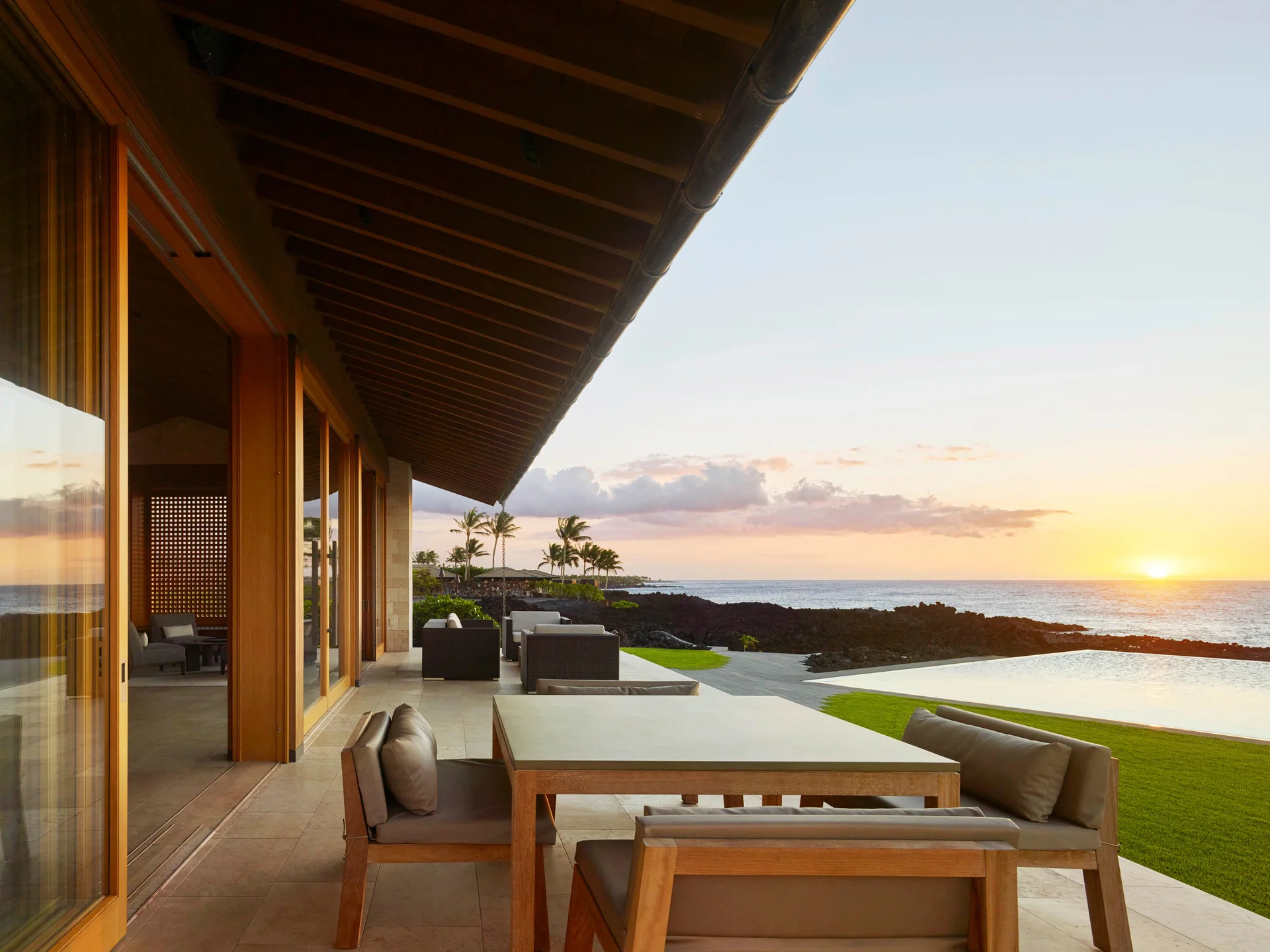 A covered outdoor patio features a wooden dining set, stone tile flooring, and an infinity pool overlooking a coastline at sunset.