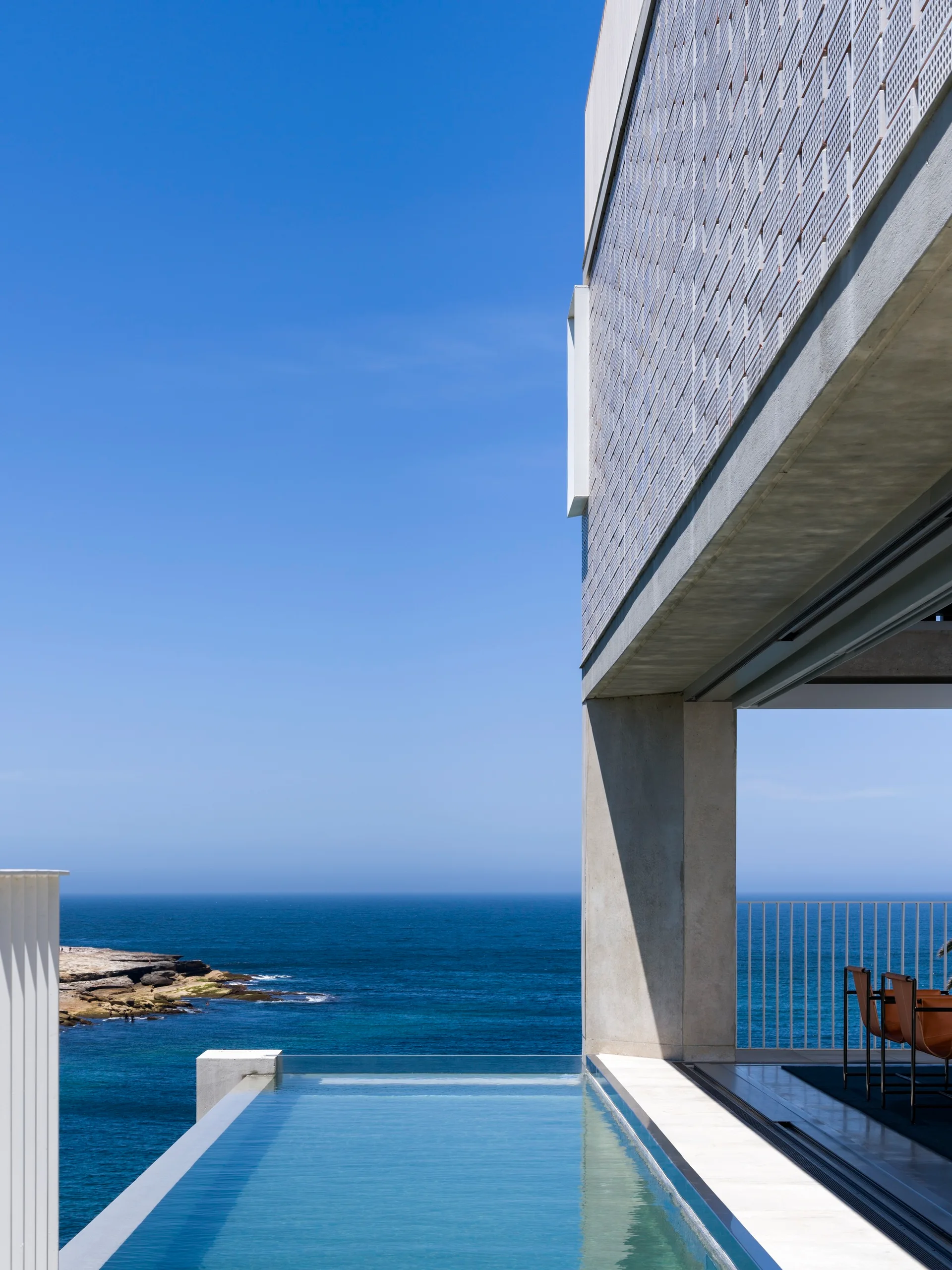 A concrete architectural structure features an infinity pool overlooking a clear blue ocean, with a covered outdoor seating area visible on the right.