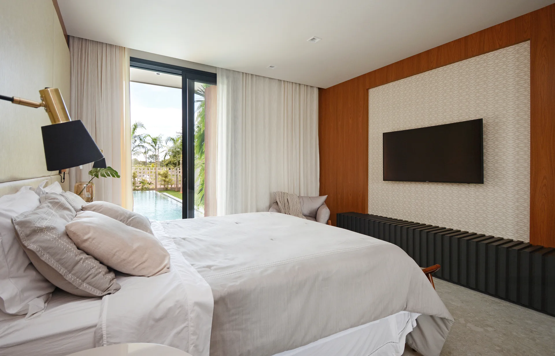 A contemporary bedroom features a neutral bed, a wall-mounted television over a dark slatted console, and sliding doors opening to a pool.