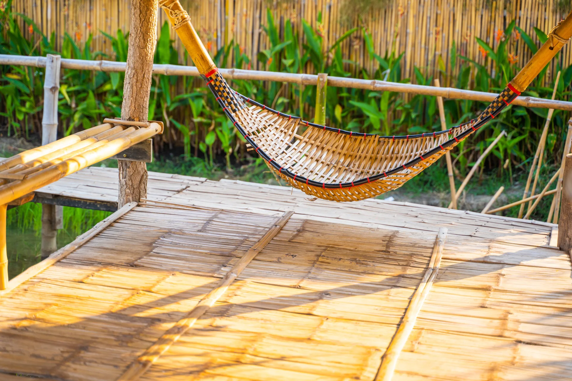 A curved, woven bamboo hammock is suspended above a slatted bamboo floor on a sunny patio with a green garden background.