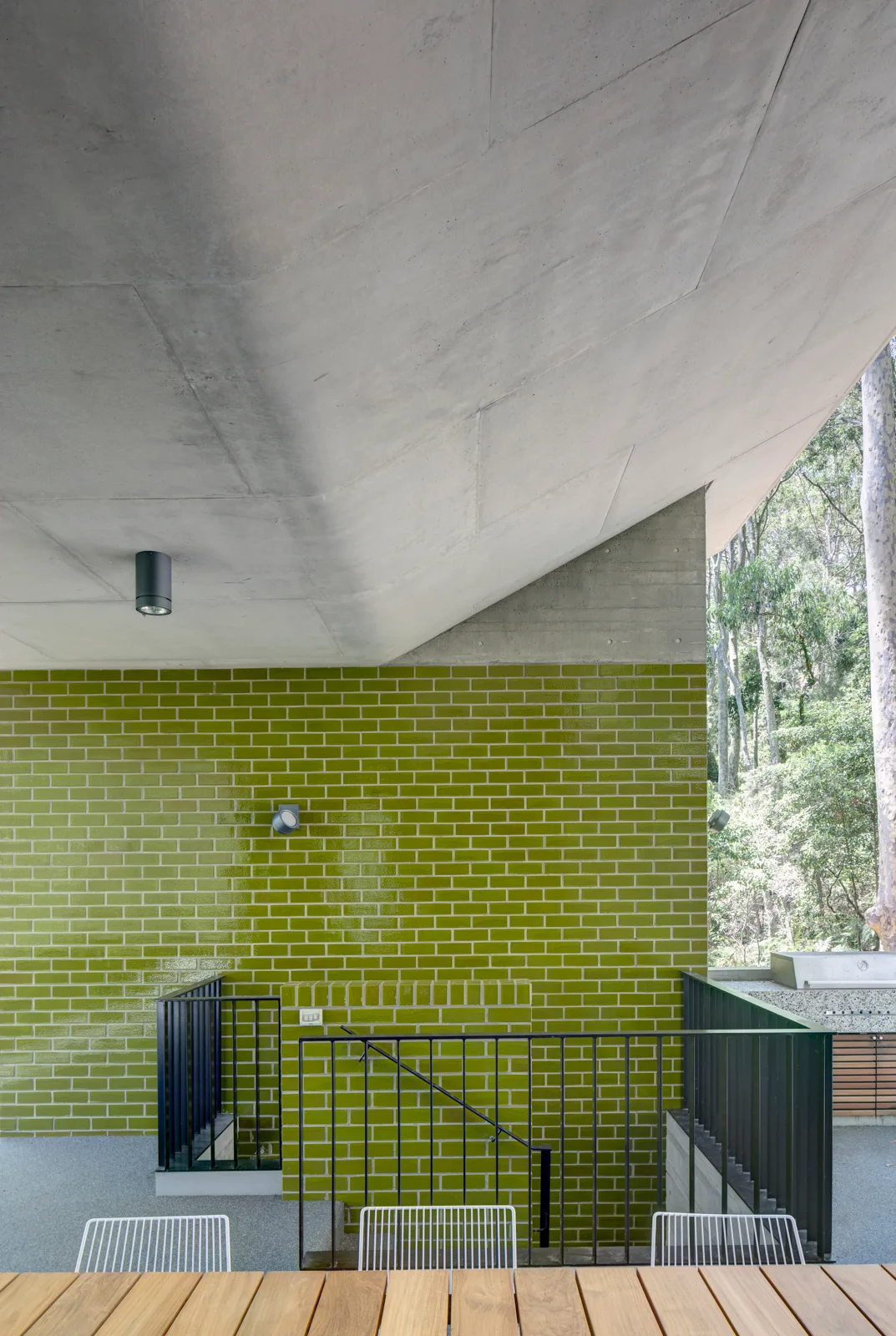 An outdoor patio features a glossy olive green brick wall, a raw concrete ceiling canopy, and a wooden dining table with wire chairs.