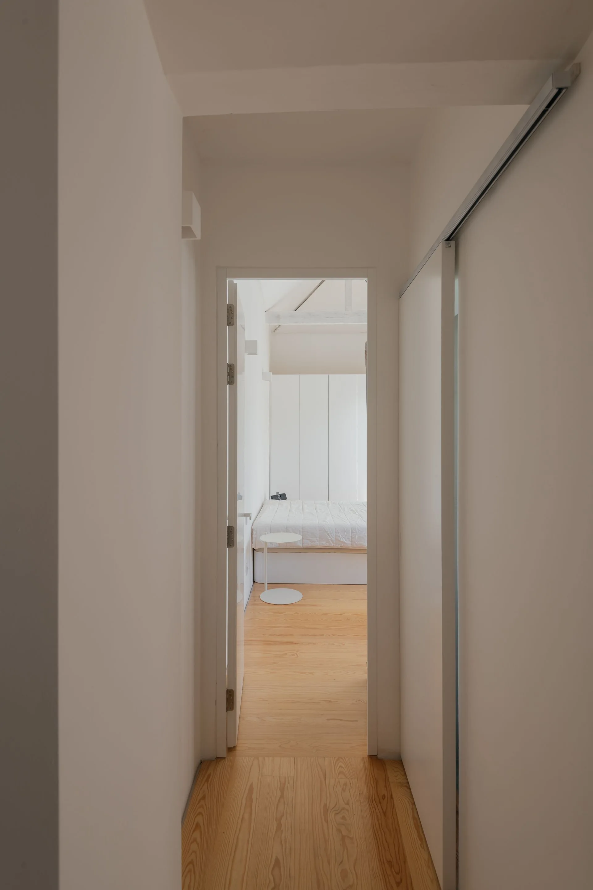 A view through a white doorframe into a minimalist bedroom with light wood floors, a low platform bed, and a small round white side table.