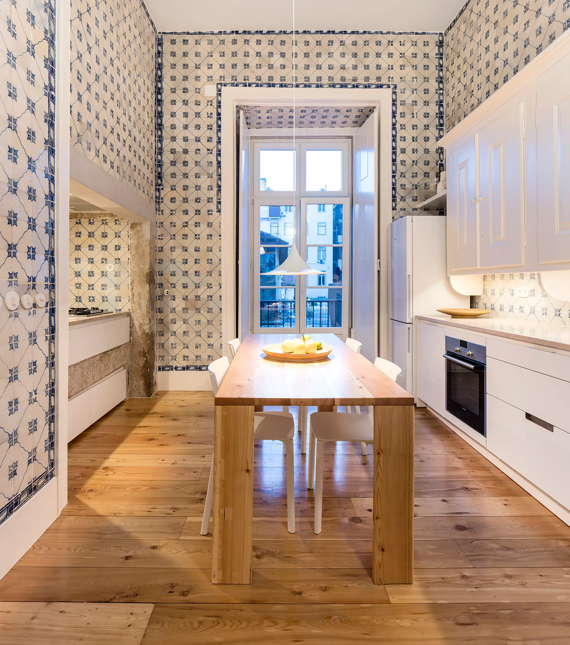 A dining kitchen featuring walls covered in blue and white patterned tiles, a central wood dining table, and modern white cabinets.