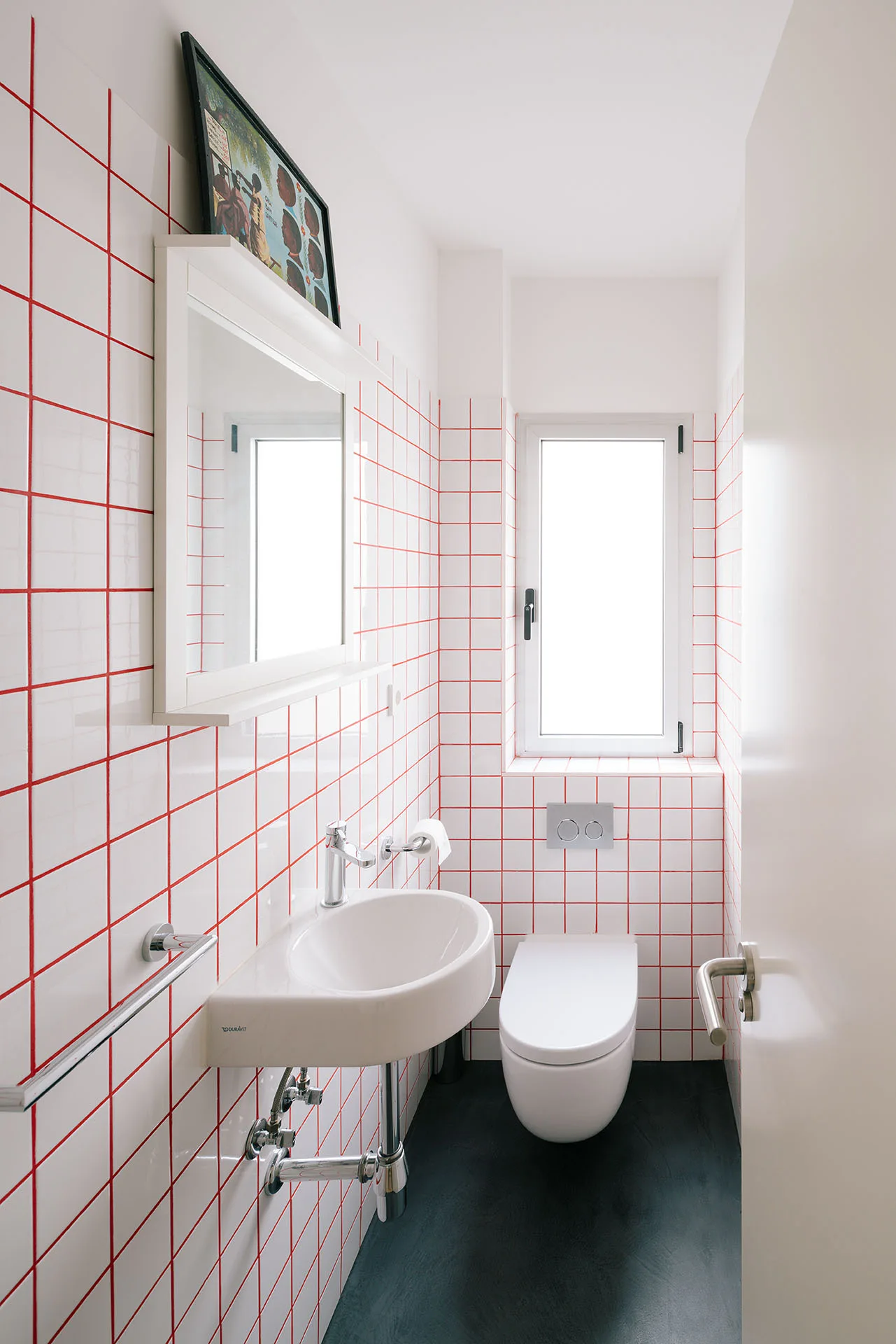 A small bathroom featuring white grid wall tiles with red grout, a wall-mounted sink, a wall-hung toilet, and a narrow window.