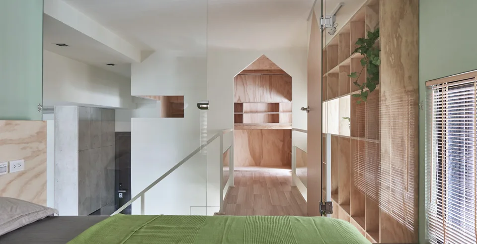 A minimalist loft bedroom features a bed with a green blanket, a glass partition, and a gabled wooden doorway leading to a plywood storage area.