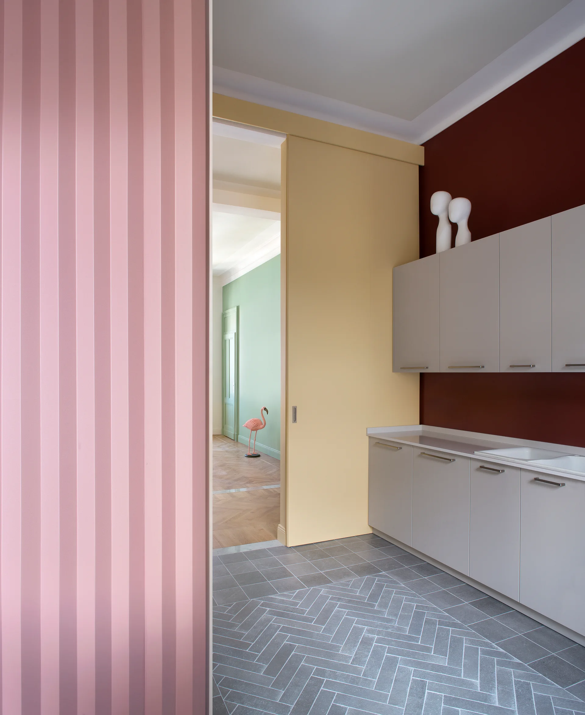 A kitchen features grey cabinets against a red wall, a yellow sliding door, pink fluted paneling, and grey herringbone tile flooring.