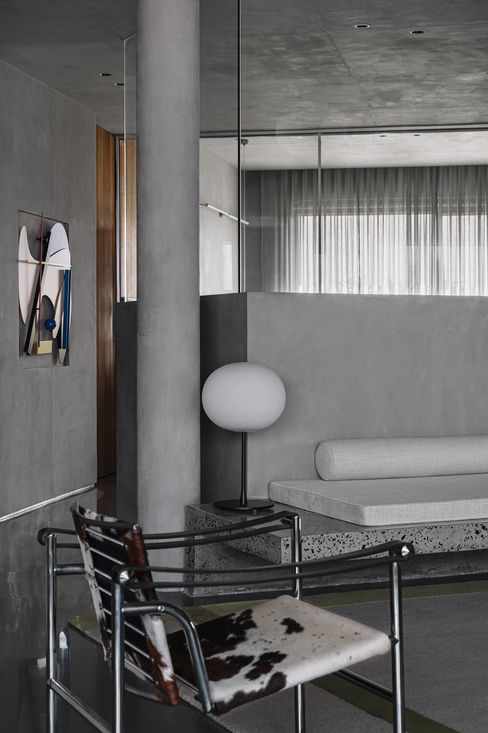 A minimalist concrete living room features a cowhide leather lounge chair, a built-in bench with a globe lamp, and an architectural pillar.