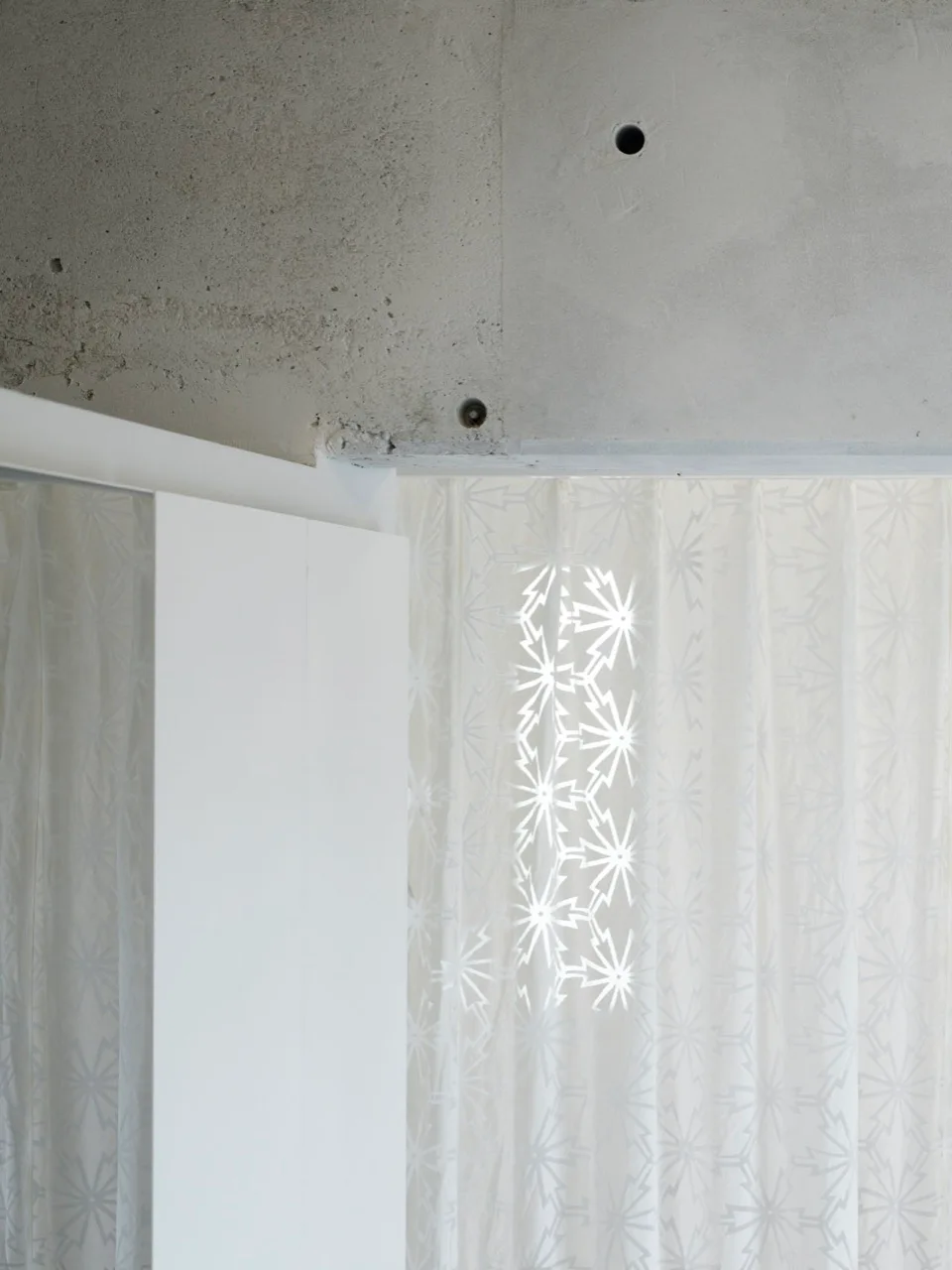 A close-up of a raw gray concrete ceiling above a sheer white curtain decorated with geometric star patterns next to a smooth white panel.