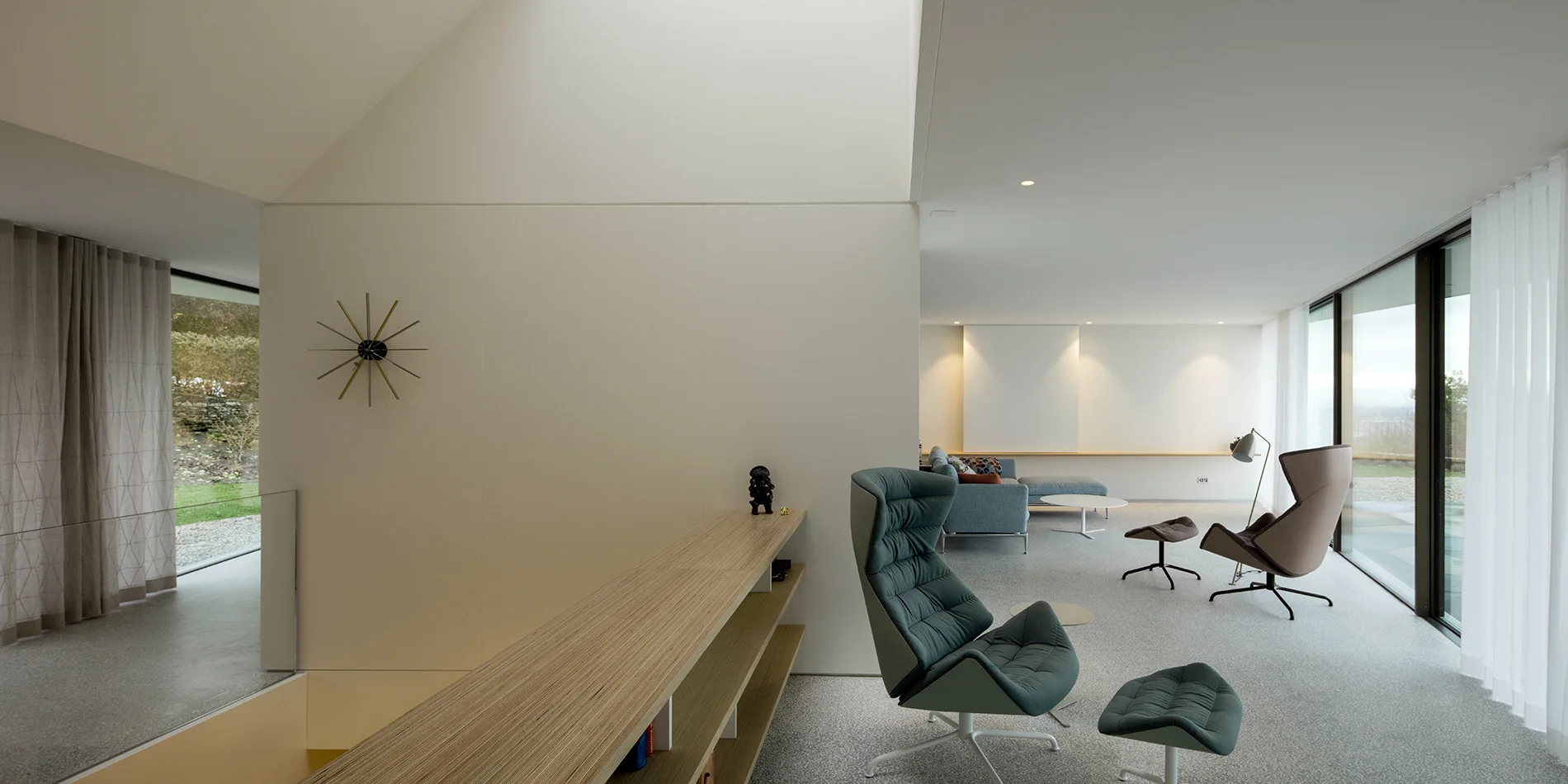 A minimalist living room features modern upholstered armchairs, a low wood shelf, and a skylight casting natural light on grey carpet.