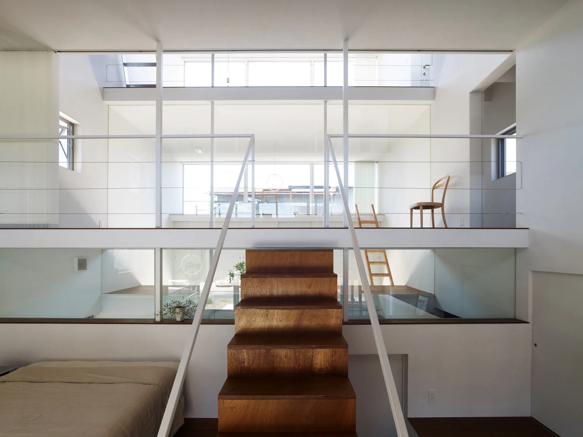 A minimalist multi-level home interior featuring a central wooden staircase, white walls, thin metal railings, and a bright mezzanine level.