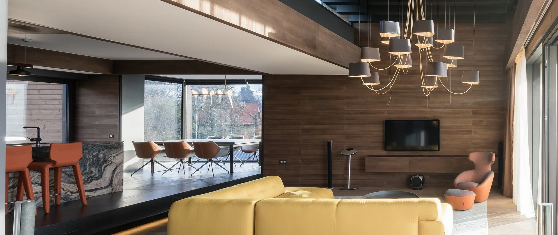 A contemporary living room featuring a bright yellow modular sofa, wood paneled walls, a large multi-shade chandelier, and a view into a dining area.