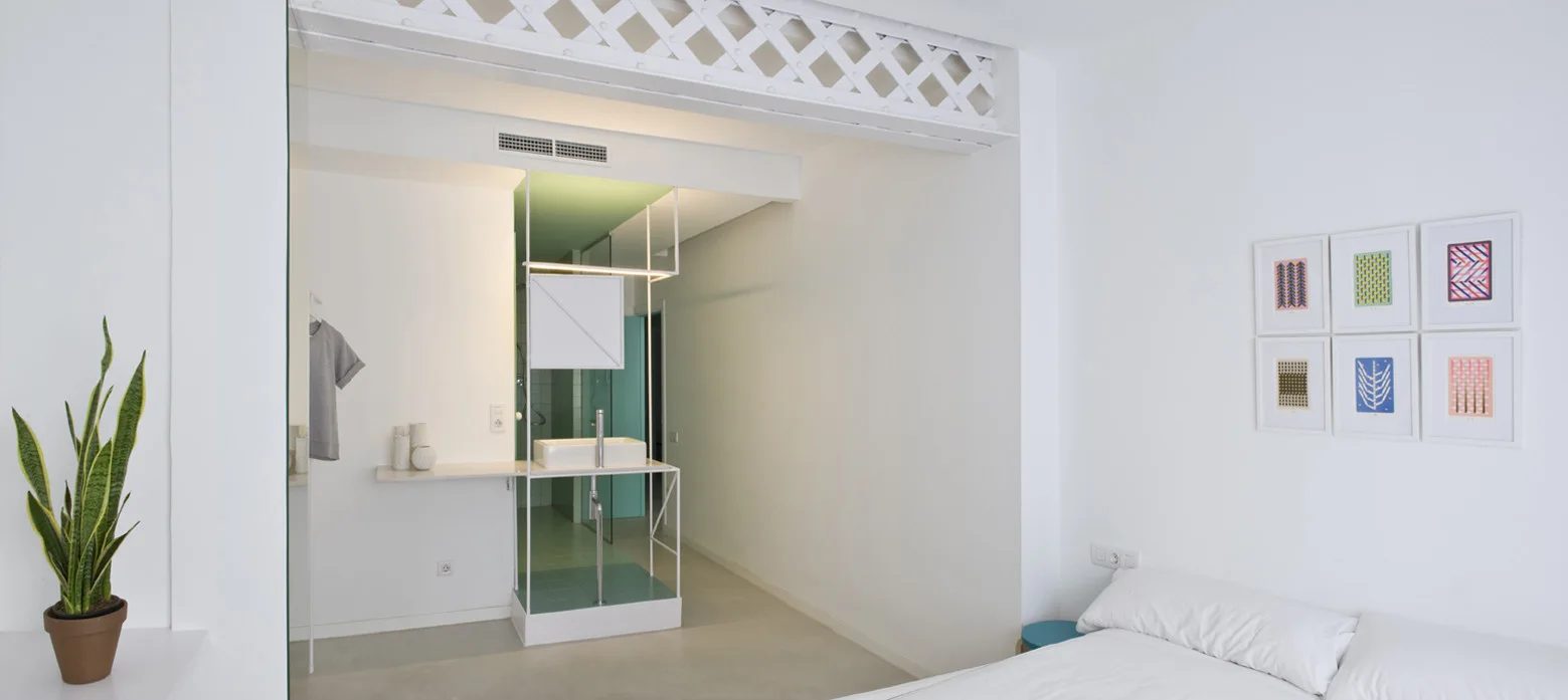 A minimalist white bedroom features a bed with white bedding, six framed art prints, and an integrated bathroom vanity with a metal frame.