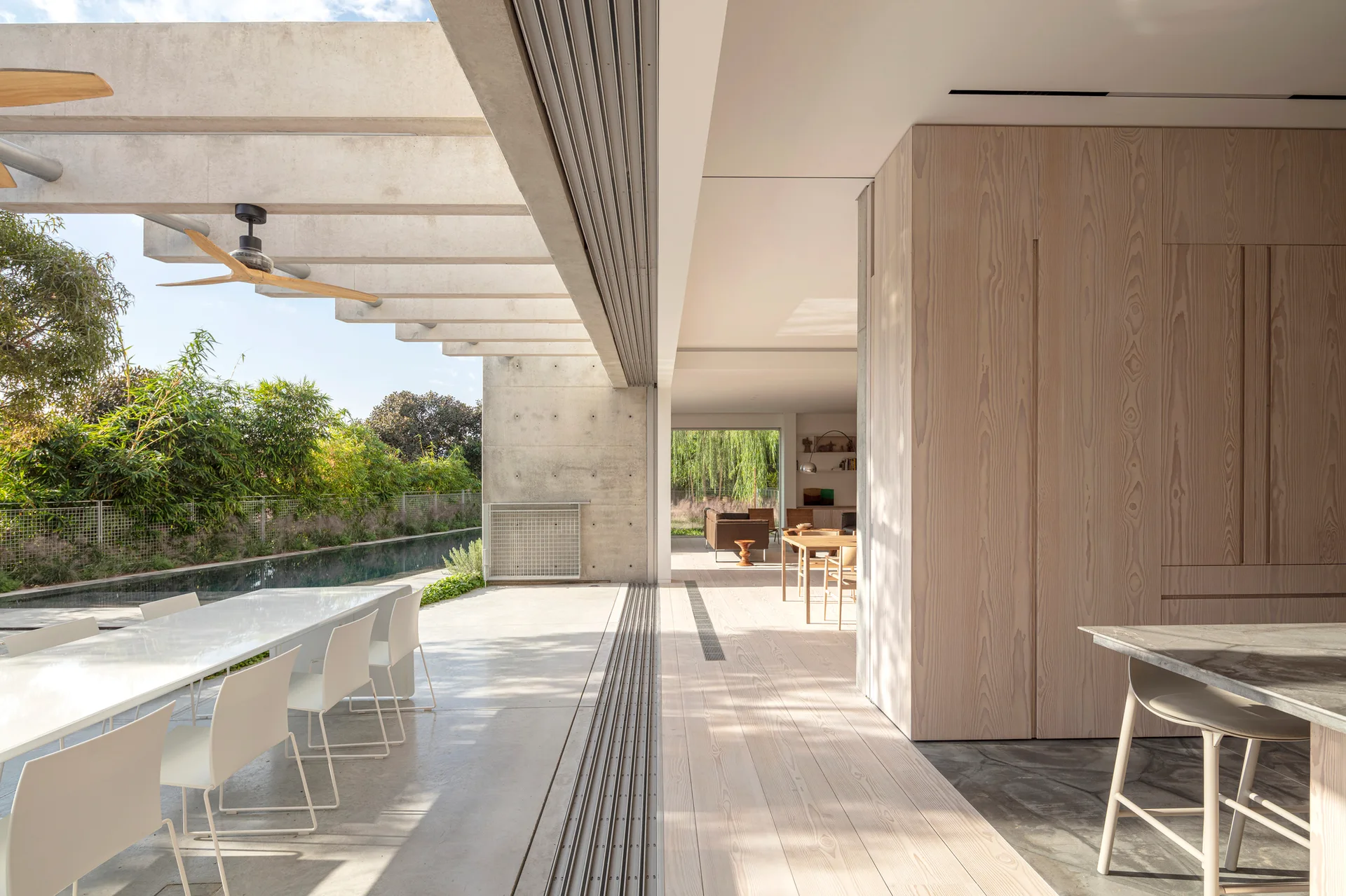 A retractable glass wall opens from a light wood kitchen to a concrete patio with a white dining table next to a swimming pool.