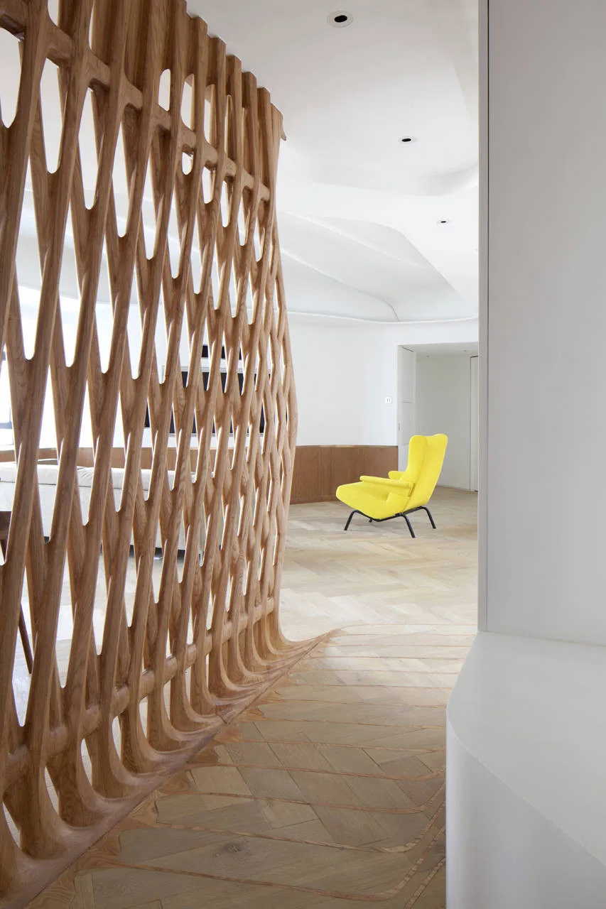 A light wood sculptural lattice screen stands on herringbone floors in a minimalist room with a bright yellow armchair in the background.