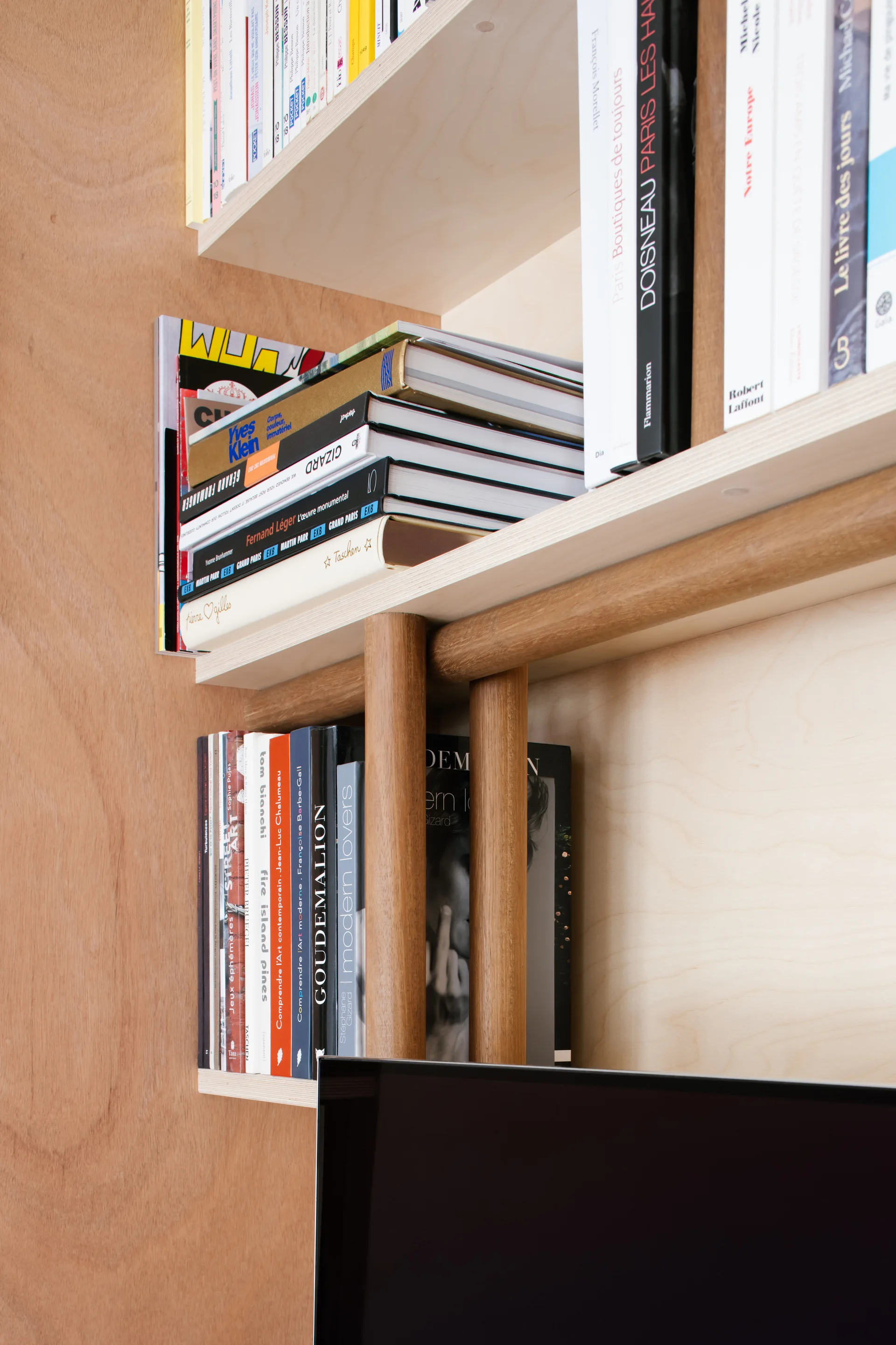 A close-up of light wood plywood shelves containing stacked books and supported by thick cylindrical wooden dowels.
