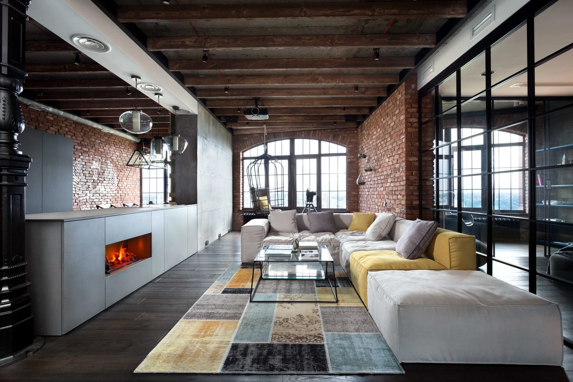 An industrial loft living room with exposed brick walls, timber ceiling beams, a modular sectional sofa, a patchwork rug, and a glass partition.