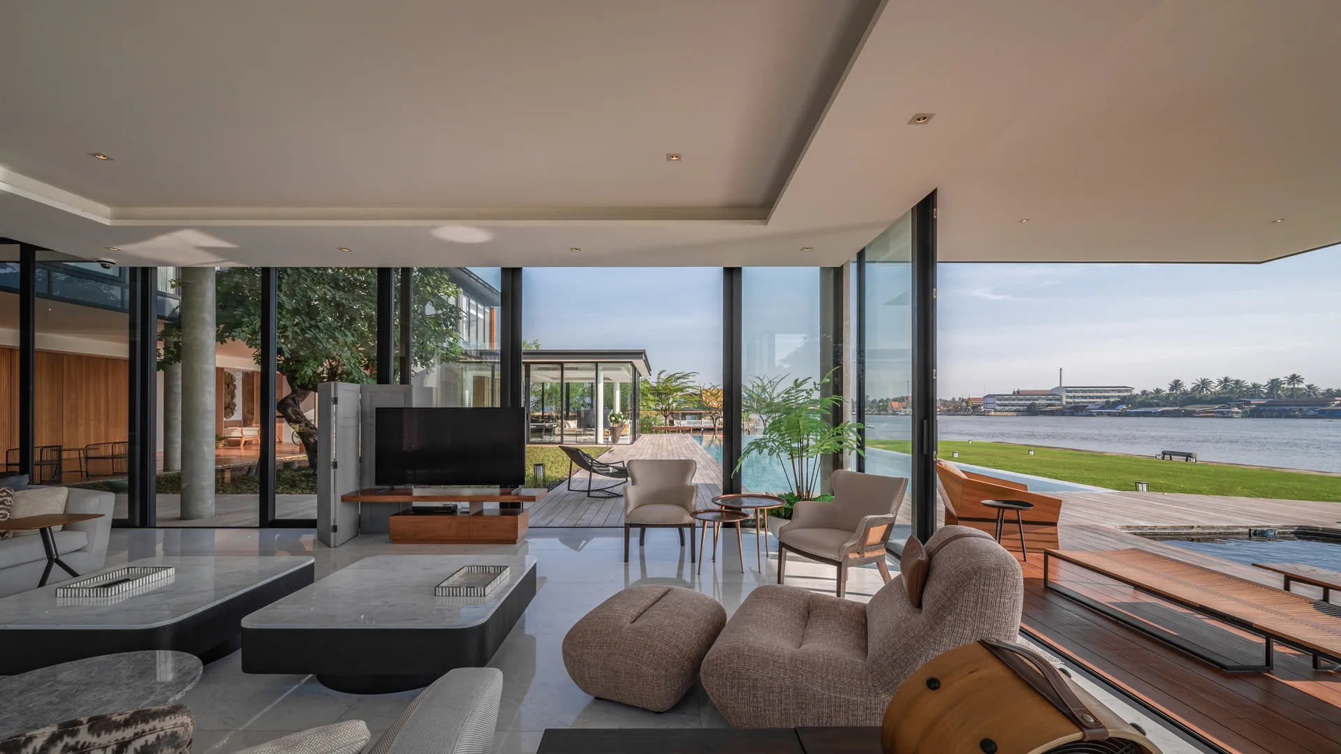 A spacious modern living room features polished marble floors, low-profile seating, and floor-to-ceiling glass walls overlooking a riverside deck.