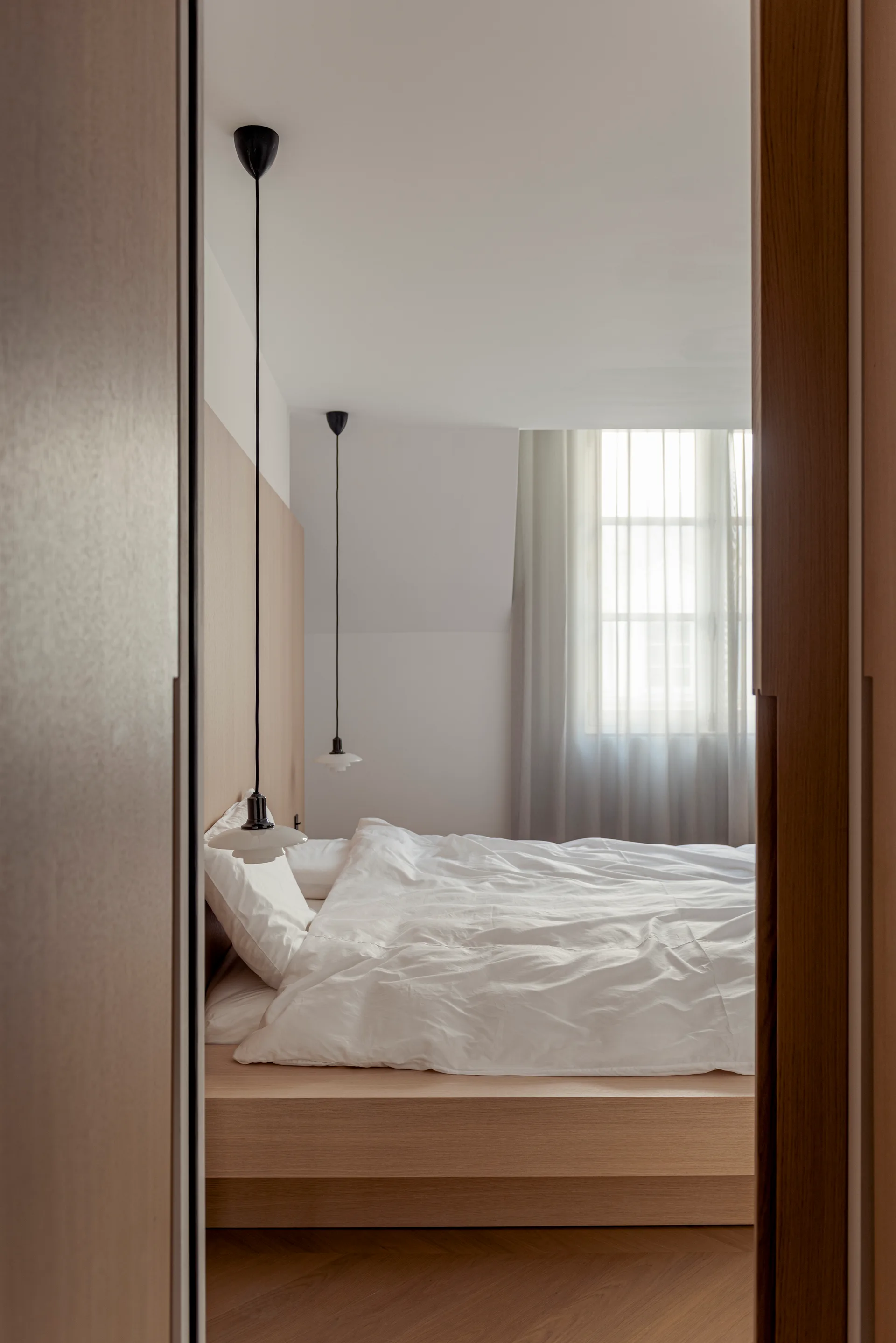 A minimalist bedroom viewed through a doorway features a light wood platform bed, matching wall paneling, and two hanging pendant lights.