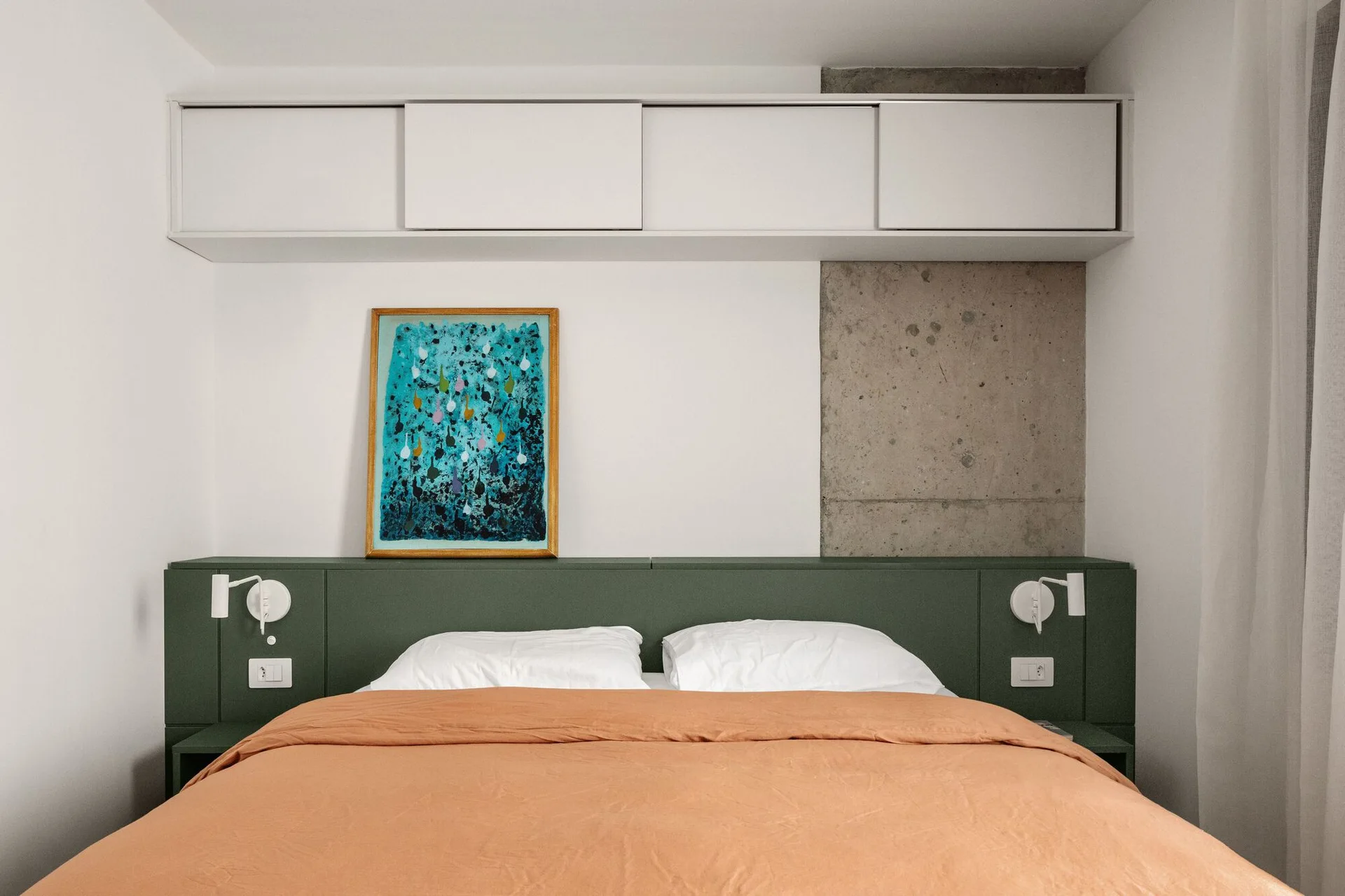A modern bedroom features a bed with an orange duvet, an integrated olive green headboard, white overhead cabinets, and an exposed concrete wall accent.