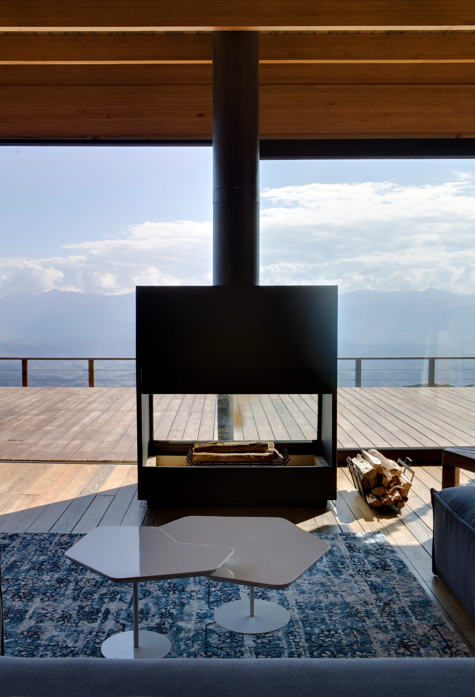 A modern living room features a central black metal fireplace in front of a glass wall that looks out onto a wooden deck and mountains.