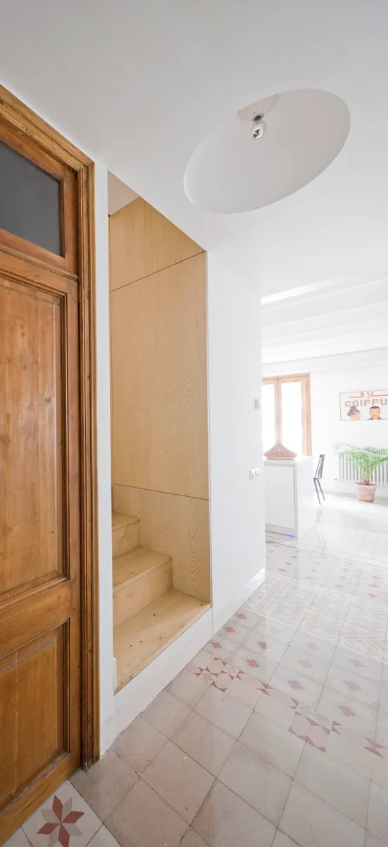 A bright hallway features an integrated light wood staircase, a rustic wooden door, an oval ceiling recess, and patterned geometric floor tiles.