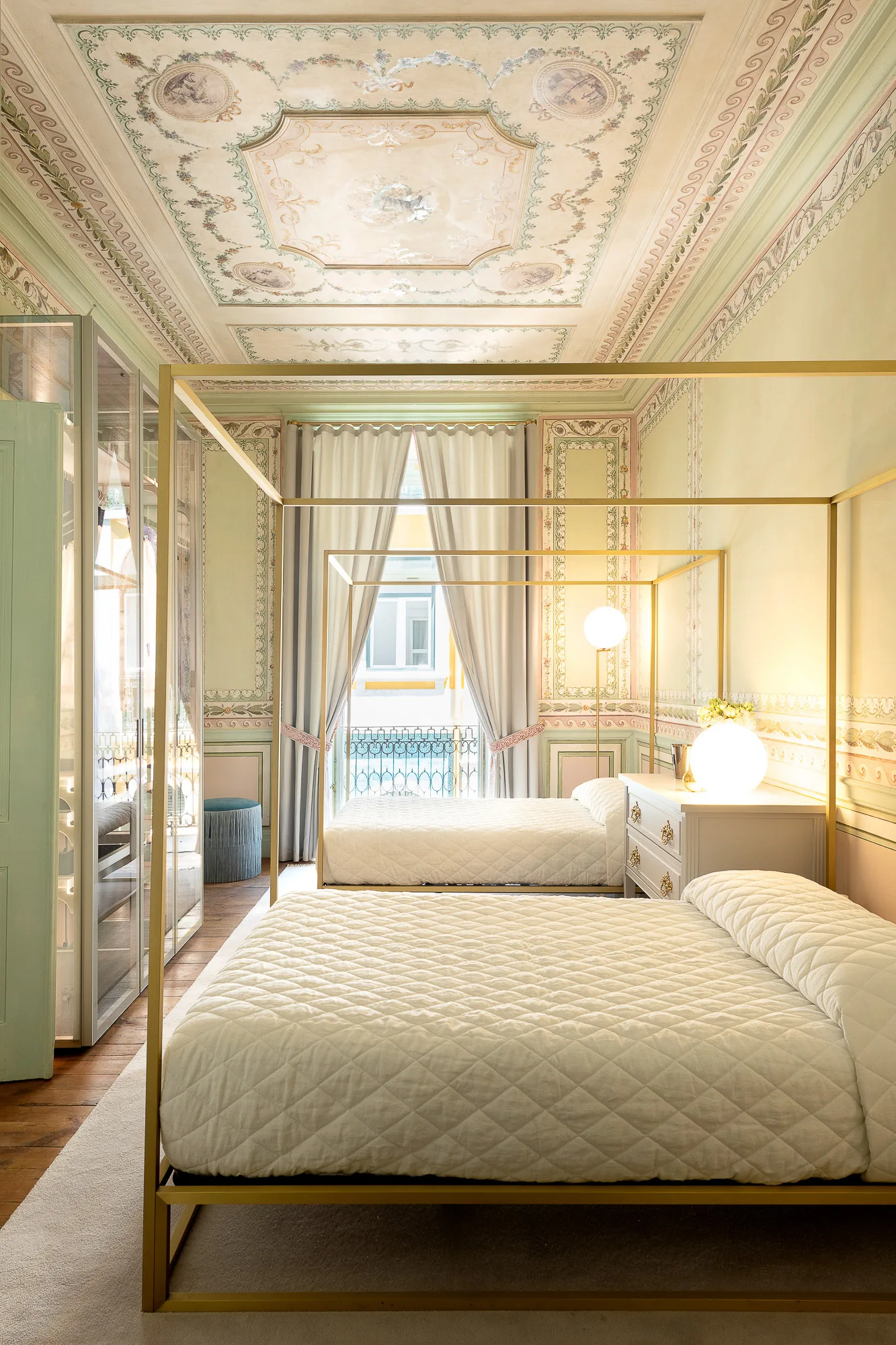 Two brass canopy beds with quilted cream covers sit inside a pale green bedroom below an ornate painted mural ceiling.