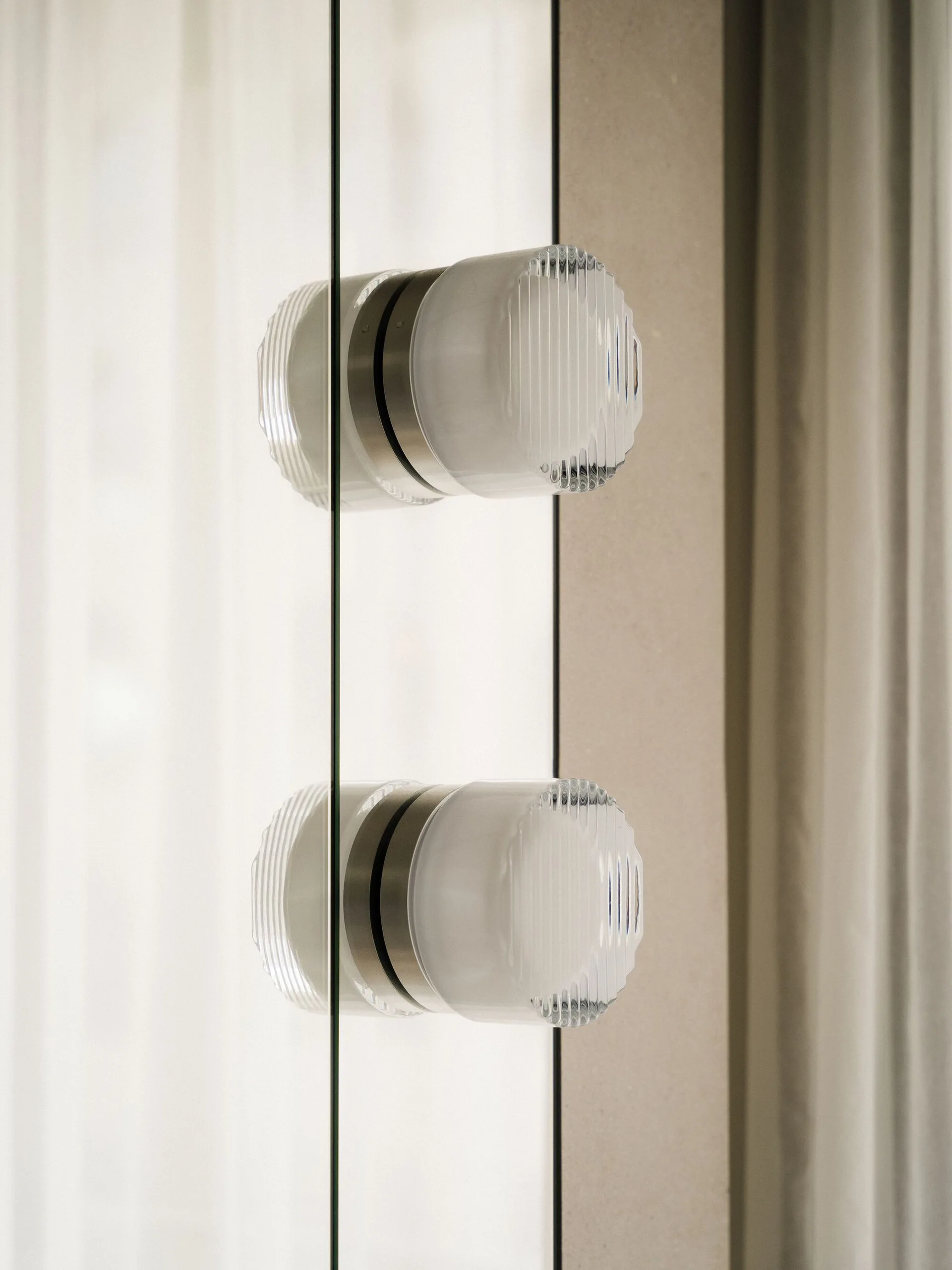 Two circular, fluted glass knobs with brushed silver metal collars are mounted vertically on a transparent glass door panel next to a beige wall.
