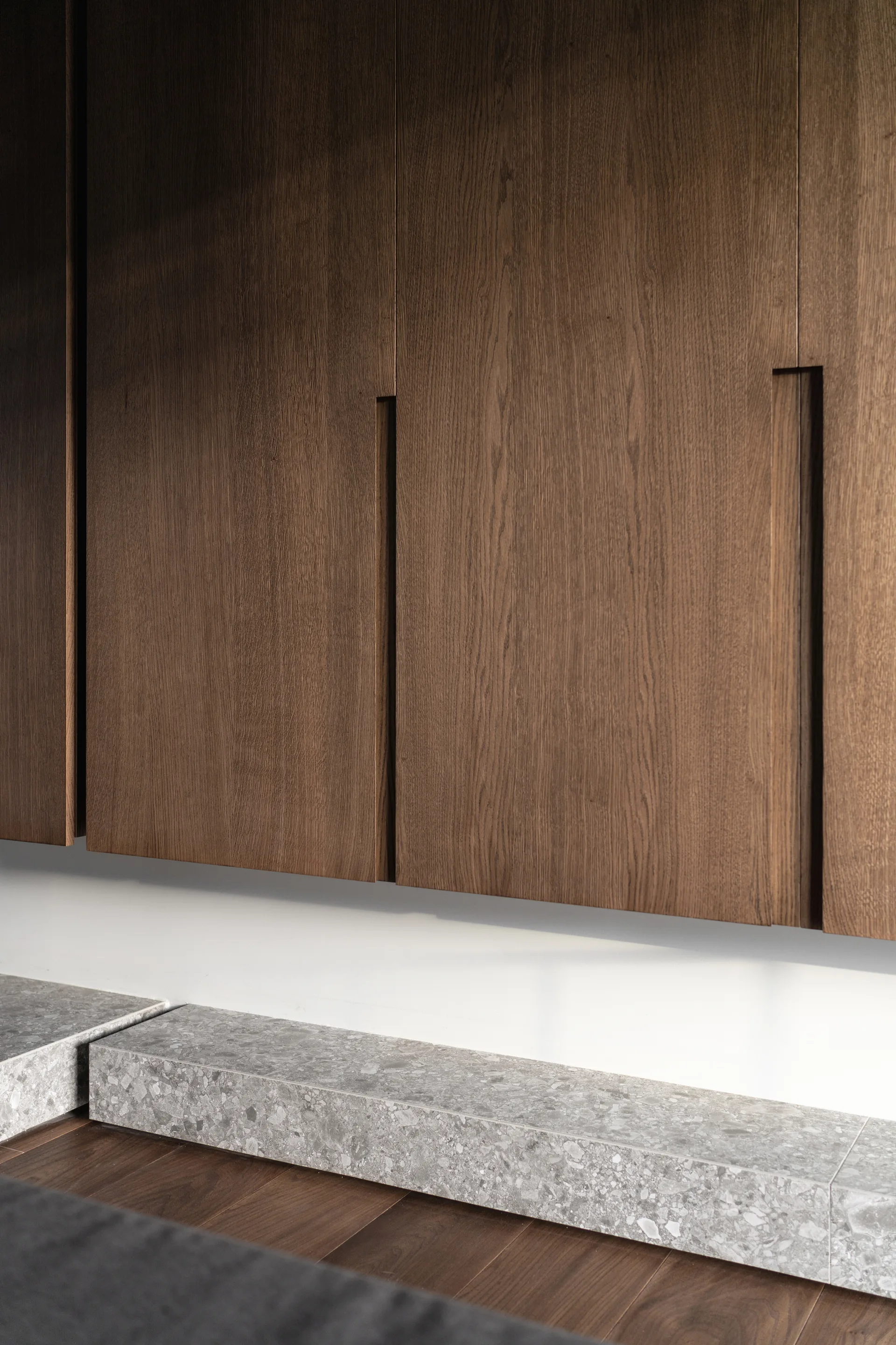 Wall-mounted dark wood cabinets with integrated vertical pulls hang above a speckled grey stone ledge and matching wood plank flooring.