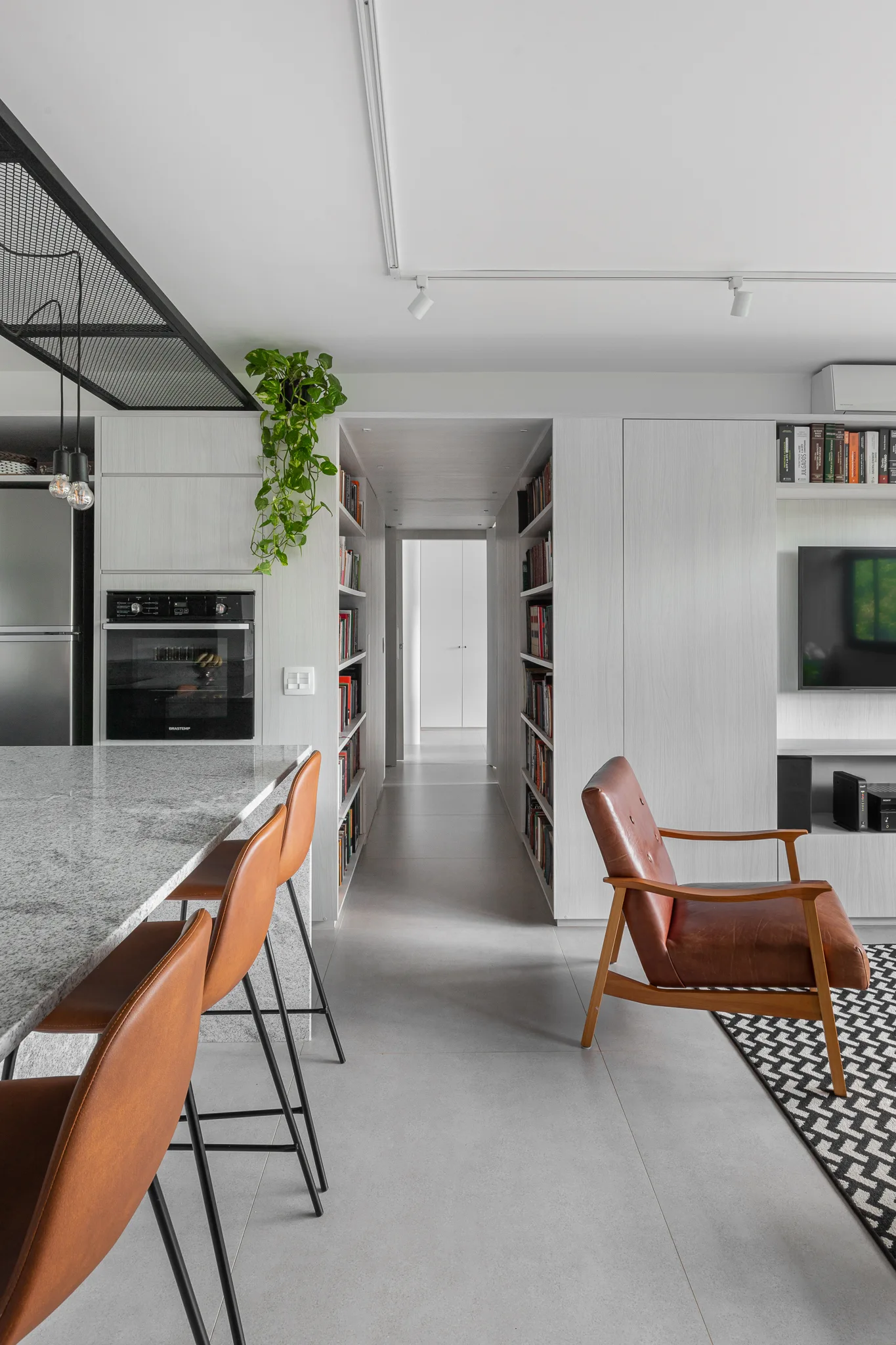 An open-concept living area and kitchen featuring a central hallway lined with built-in bookshelves, a leather armchair, and leather bar stools.
