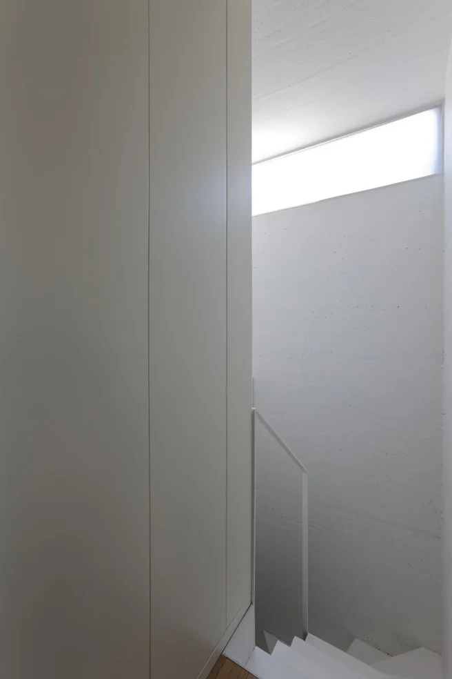 A minimalist stairwell featuring a textured concrete wall on one side and smooth white paneling on the other, lit by a high horizontal window.