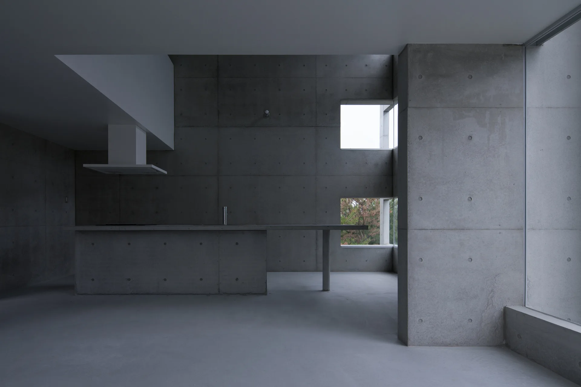 A minimalist brutalist kitchen features exposed concrete walls, a monolithic concrete island, and two stacked square windows.